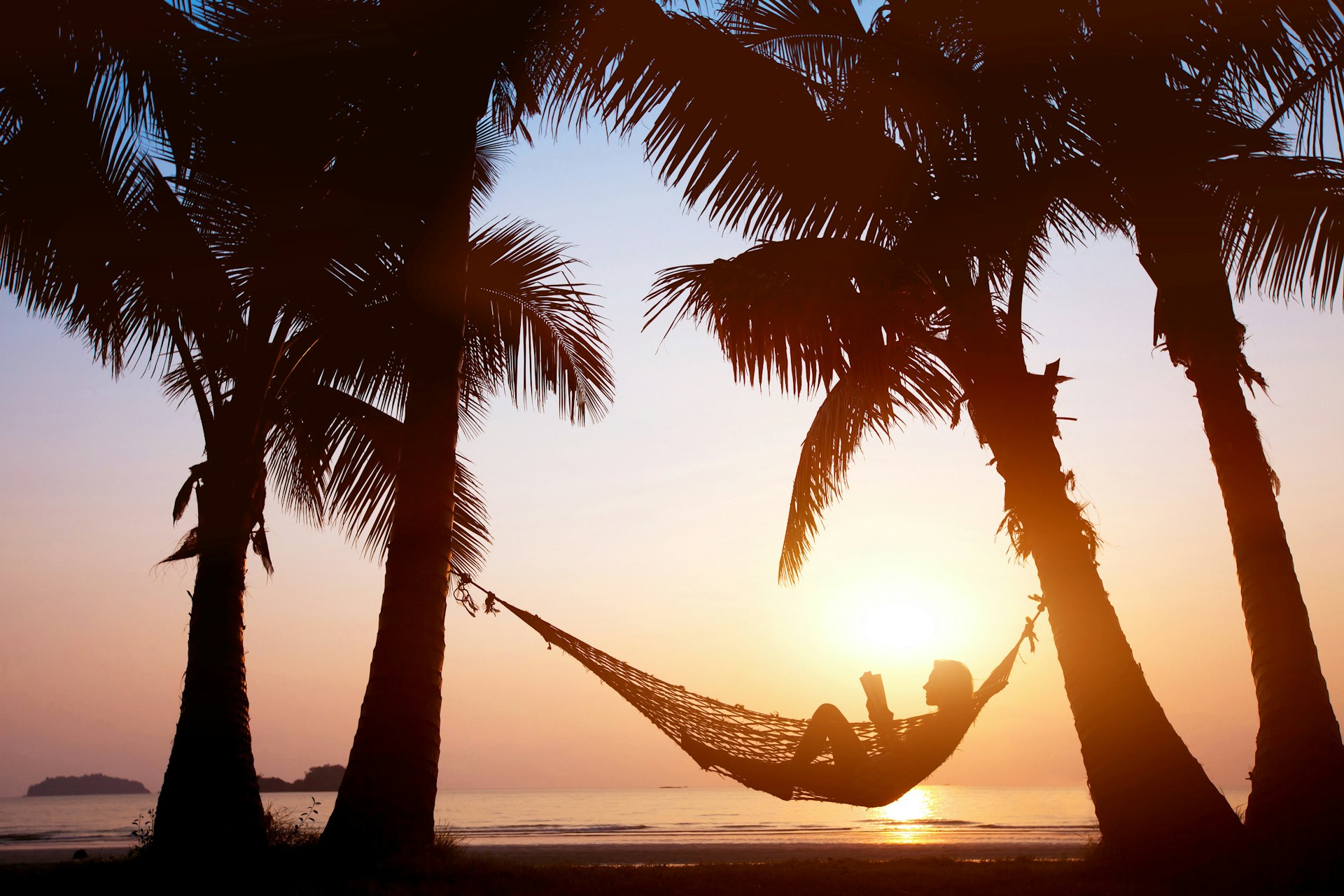 A figure relaxing in a beach hammock, backlit by the sun