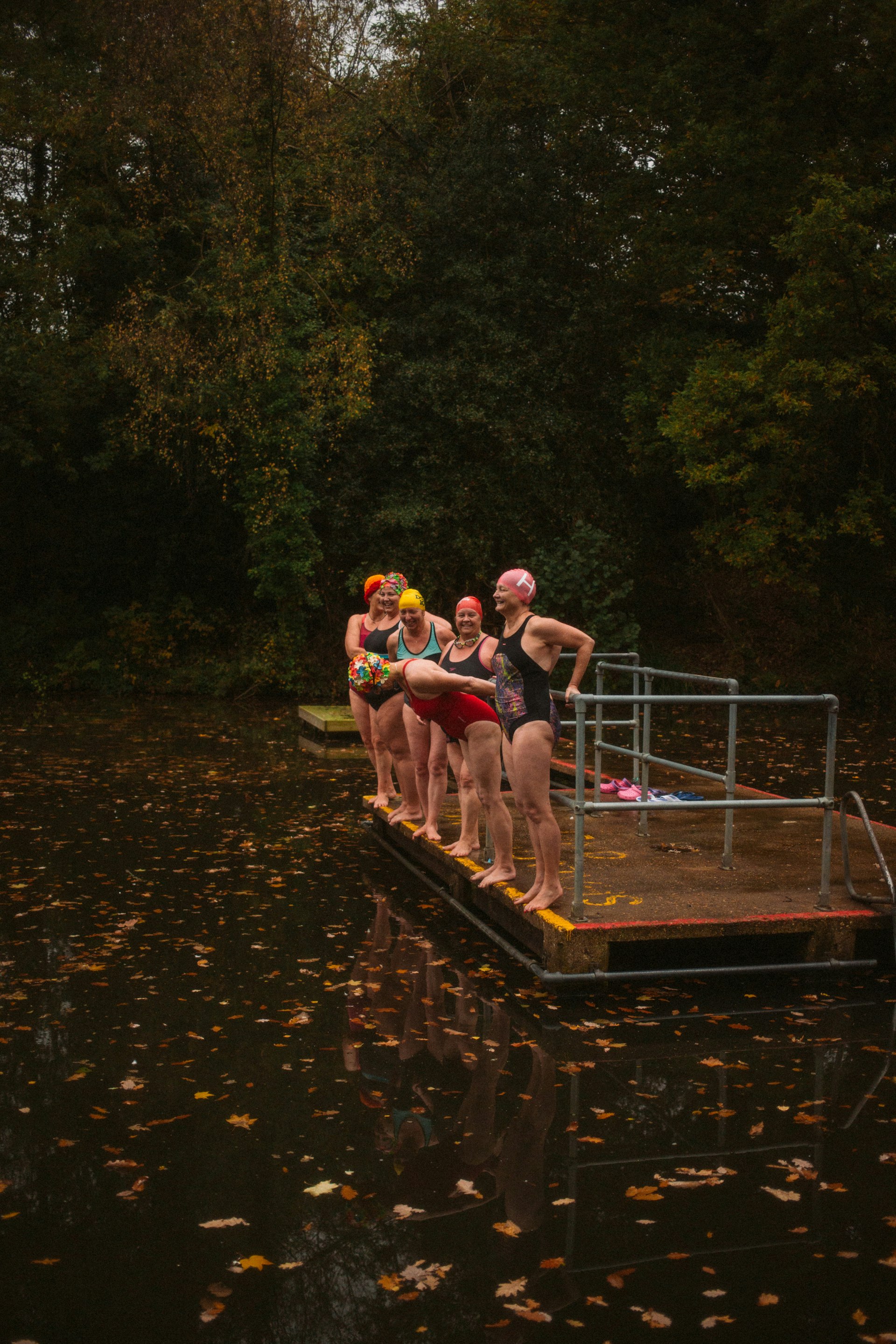 Women in swim gear stand on a platform ready to jump into a swimming pond