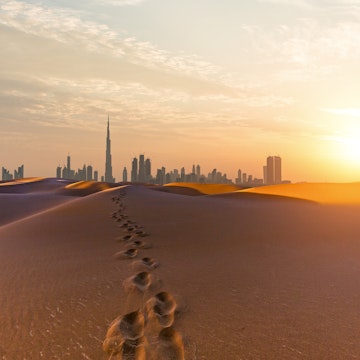 Footsteps in desert sand heading towards skyscrapers of the Dubai city skyline at dawn.