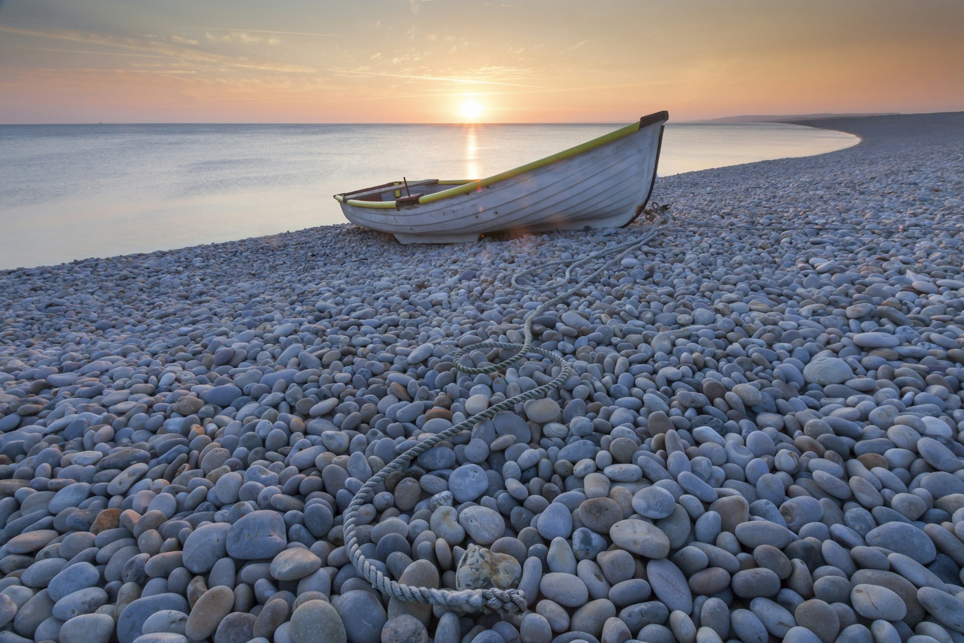 A rowing boat sits on a pebbly beach at sunset. The surface of the sea behind is extremely still and smooth