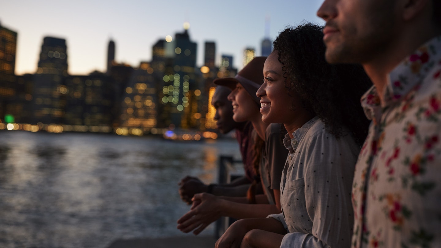Four people sitting by the river at dusk with the Manhattan skyline beyond.