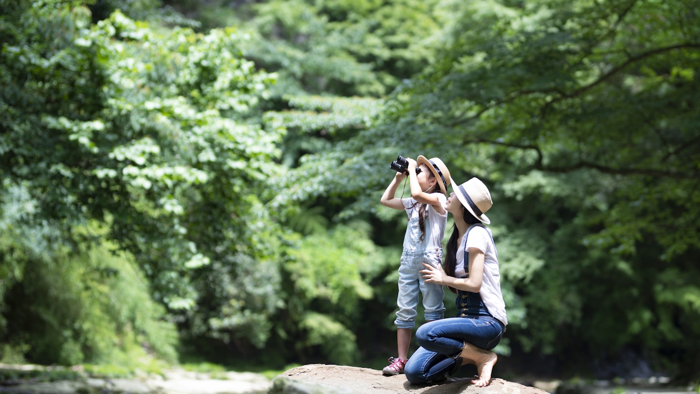 Mother kneeling beside her daughter who is looking through binoculars near a river.