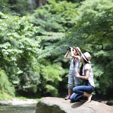 Mother kneeling beside her daughter who is looking through binoculars near a river.