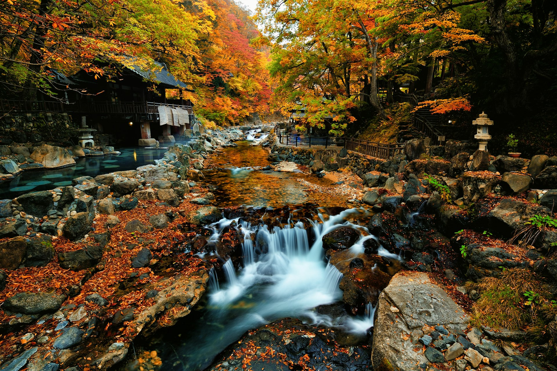 Colorful autumn leaves fill the trees and fallen ones sit on a river above a small waterfall near a hot springs in Japan
