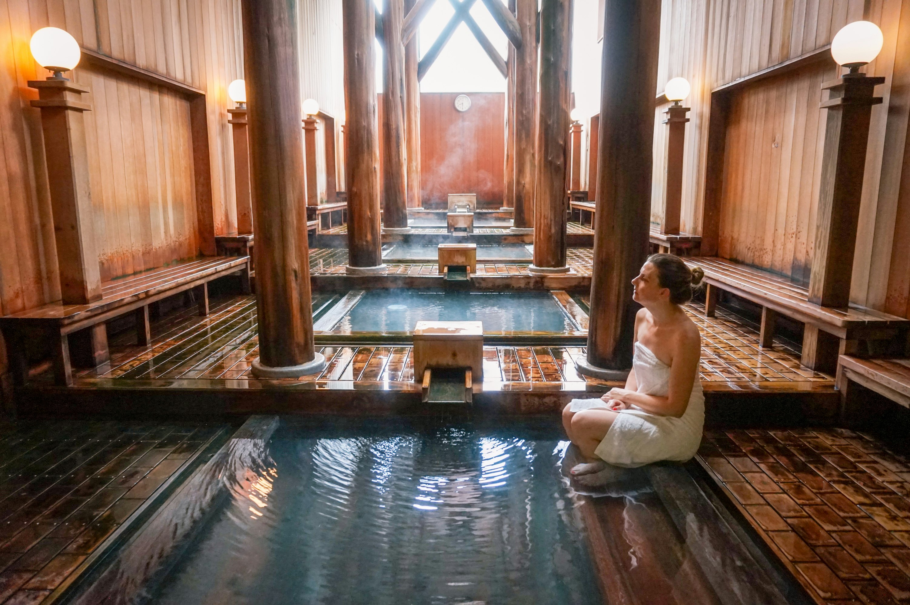 A woman wrapped in a towel sits with her feet in the water at an indoor bath surrounded by wooden walls and posts