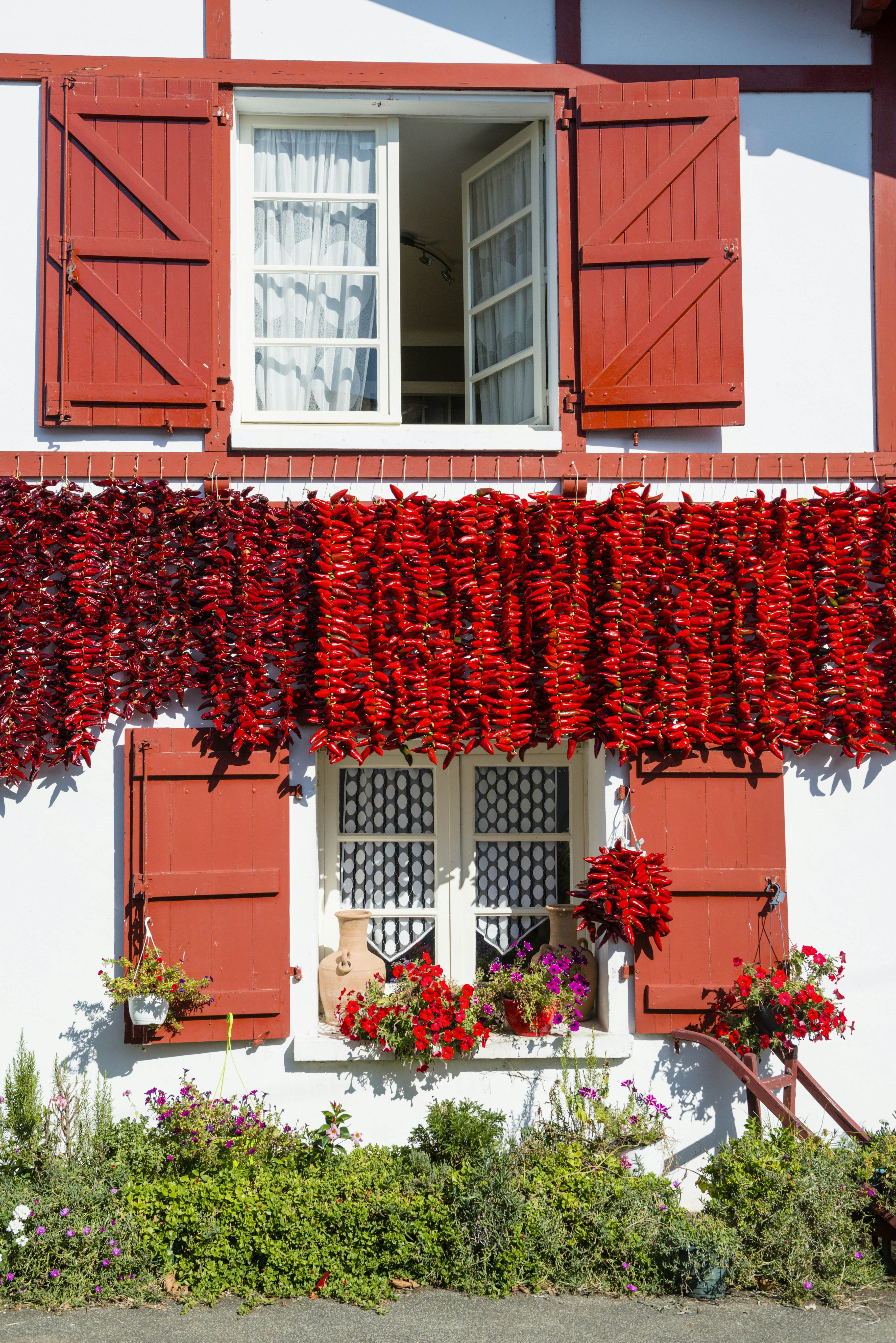A white building with red shutters around the windows. Chilli peppers hang below the upstairs window