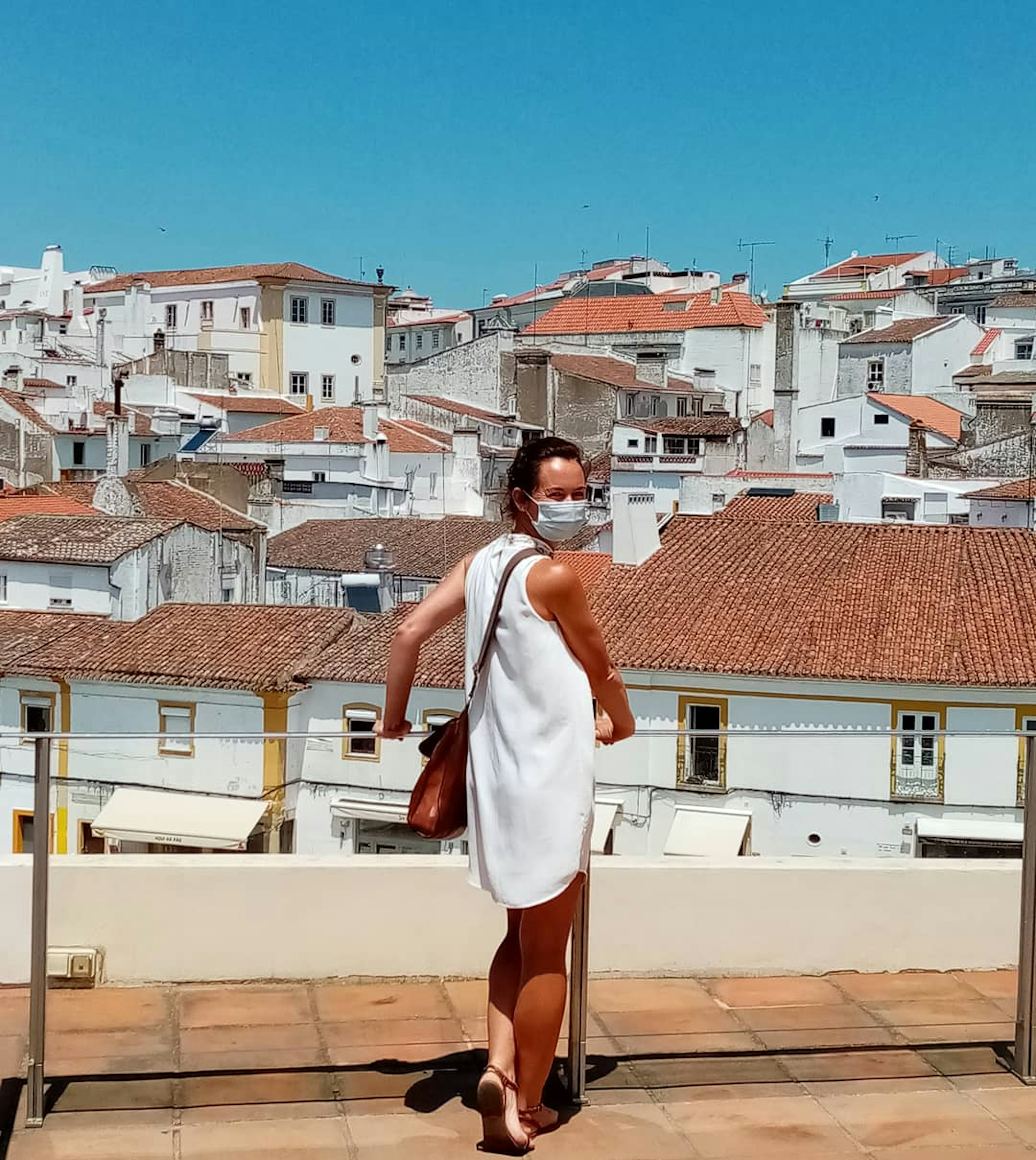 A woman stands facing a city view or red-roofed buildings. She's turning back towards the camera and wearing a face mask