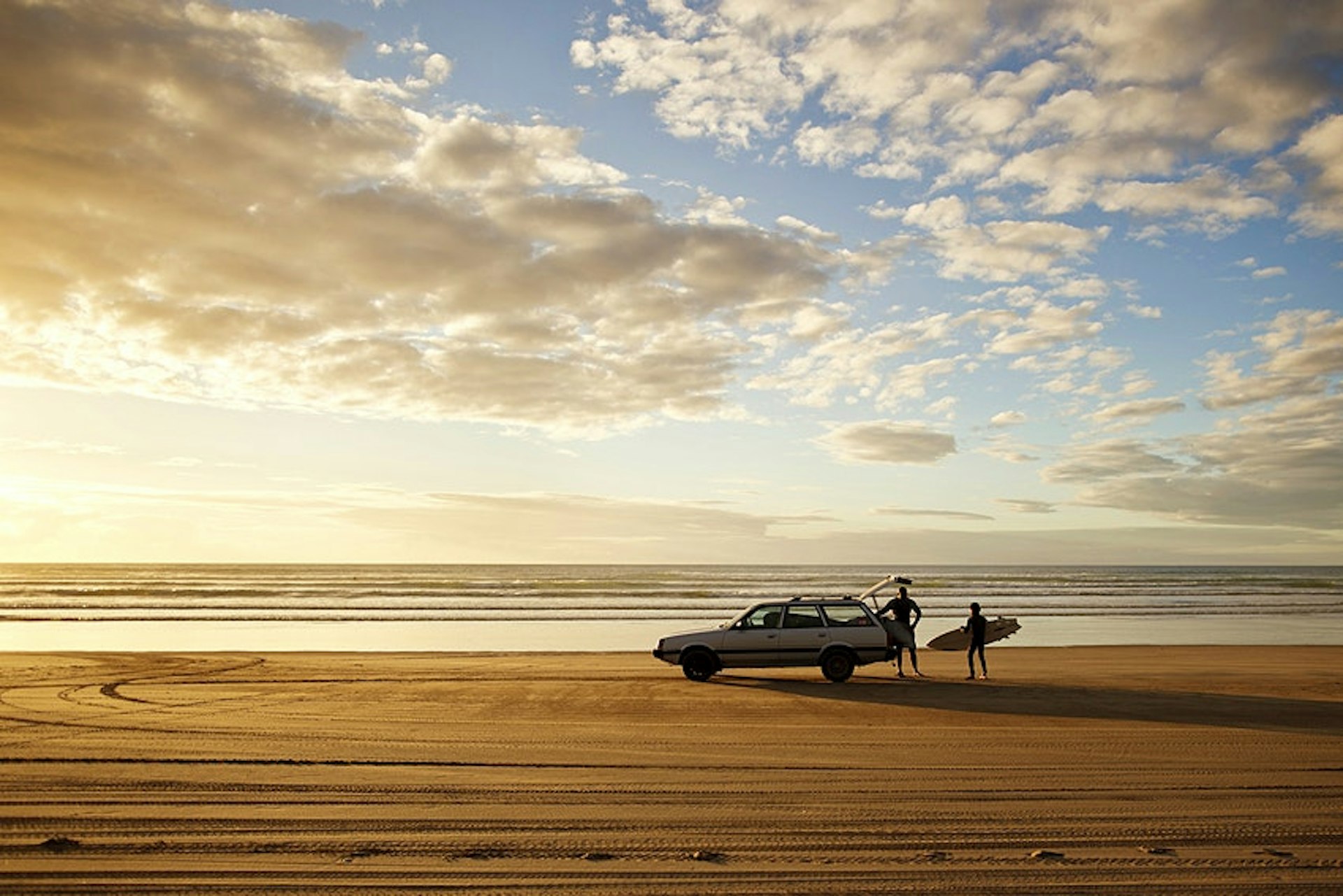 A car parked on a sandy beach as two surfers unload their boards 