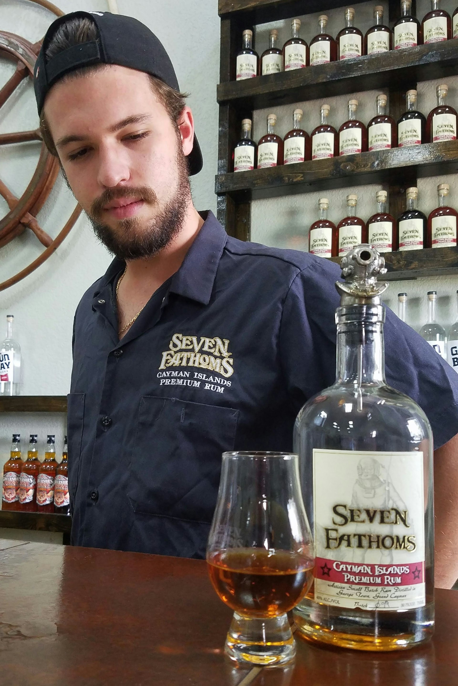 A man stands behind a bar looking and a glass and a bottle of brown-flavored rum