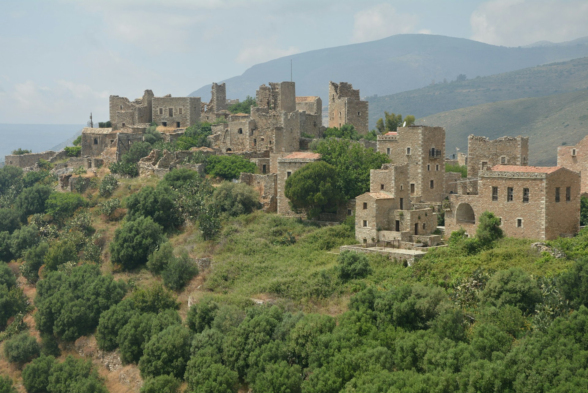 View of the stone buildings of the Vathia tower houses in the Mani region of Peloponnese. Some of the buildings are crumbling on the green mountainside. 