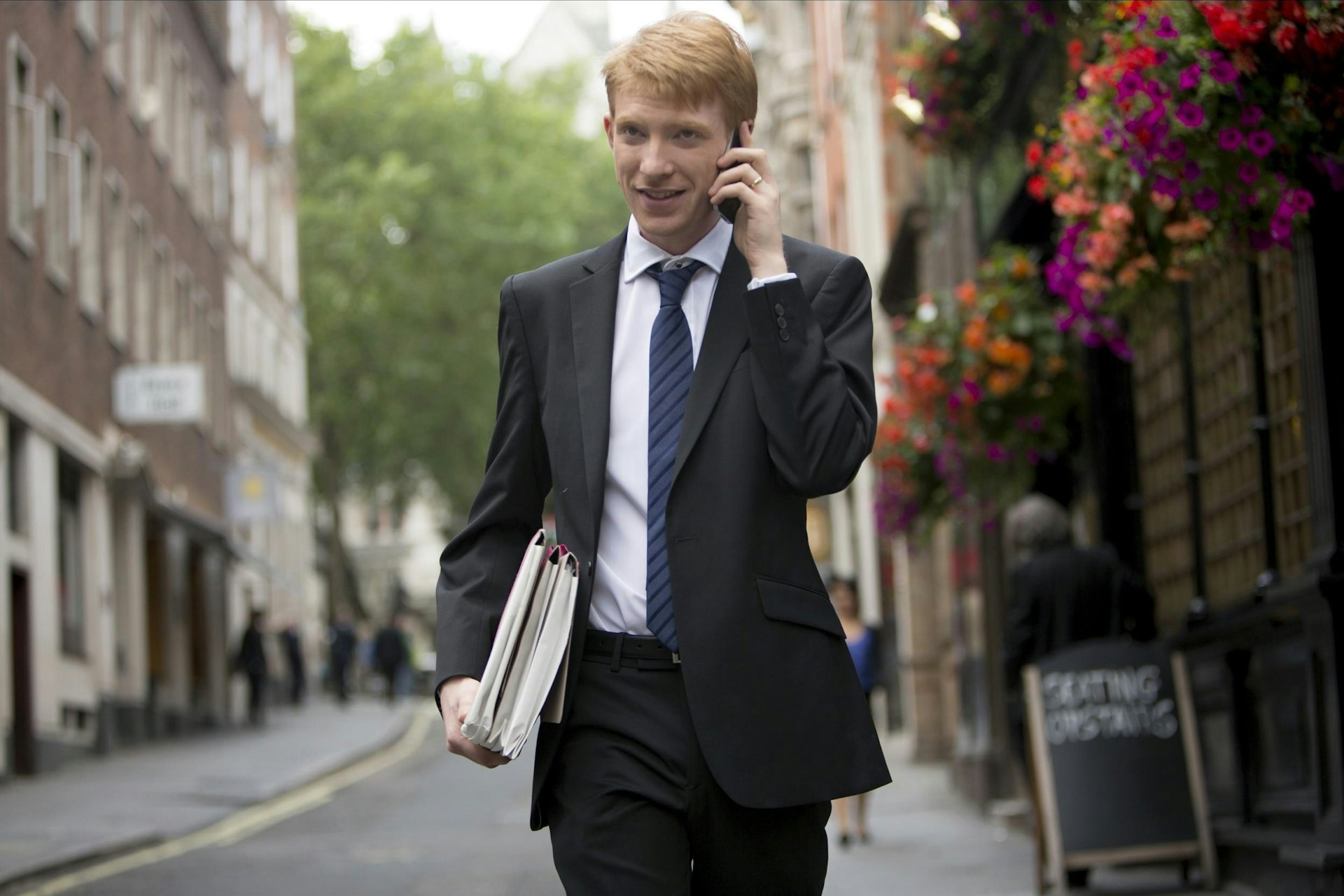 Domhnall Gleeson in About Time, walking down a cobblestone street wearing a suit with a phone in his hand. A pub with flowers is seen out of focus in the background.