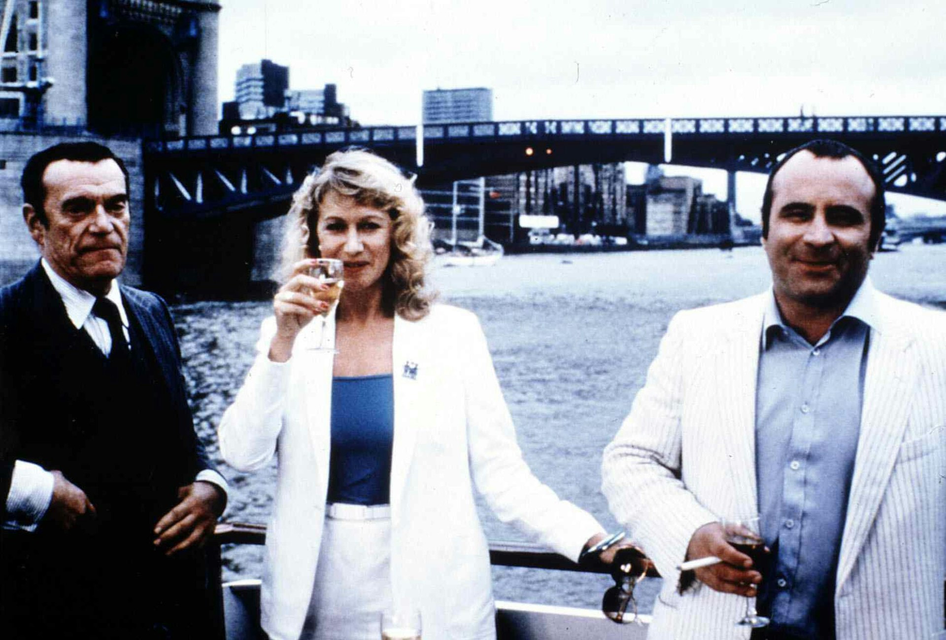 Eddie Constantine, Helen Mirren and Bob Hoskins stand on a bridge in London. Mirren is holding a glass of wine up.
