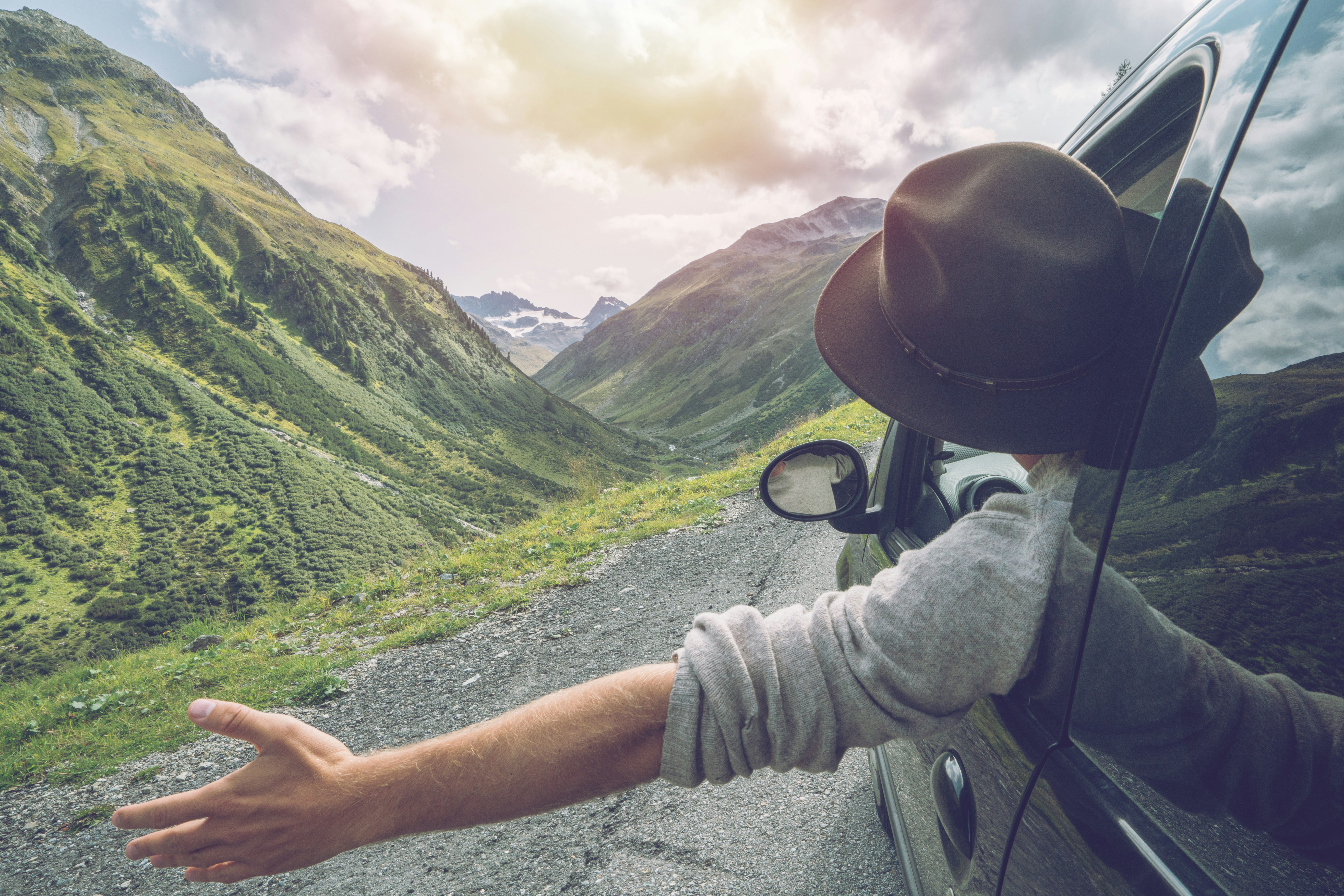 Young man in car on mountain road looks out from window car, outstretched arms for freedom. Mountain landscape in Summer, shot in Graubunden Canton, Switzerland.