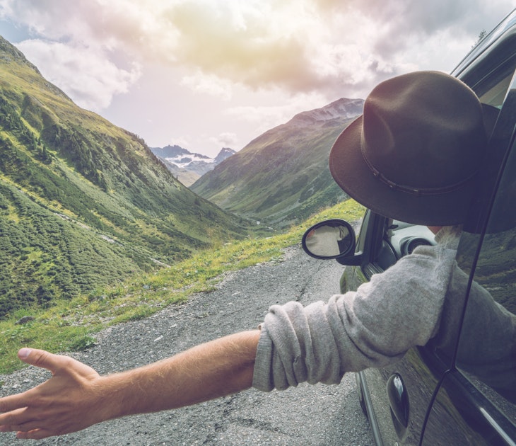 Young man in car on mountain road looks out from window car, outstretched arms for freedom. Mountain landscape in Summer, shot in Graubunden Canton, Switzerland.