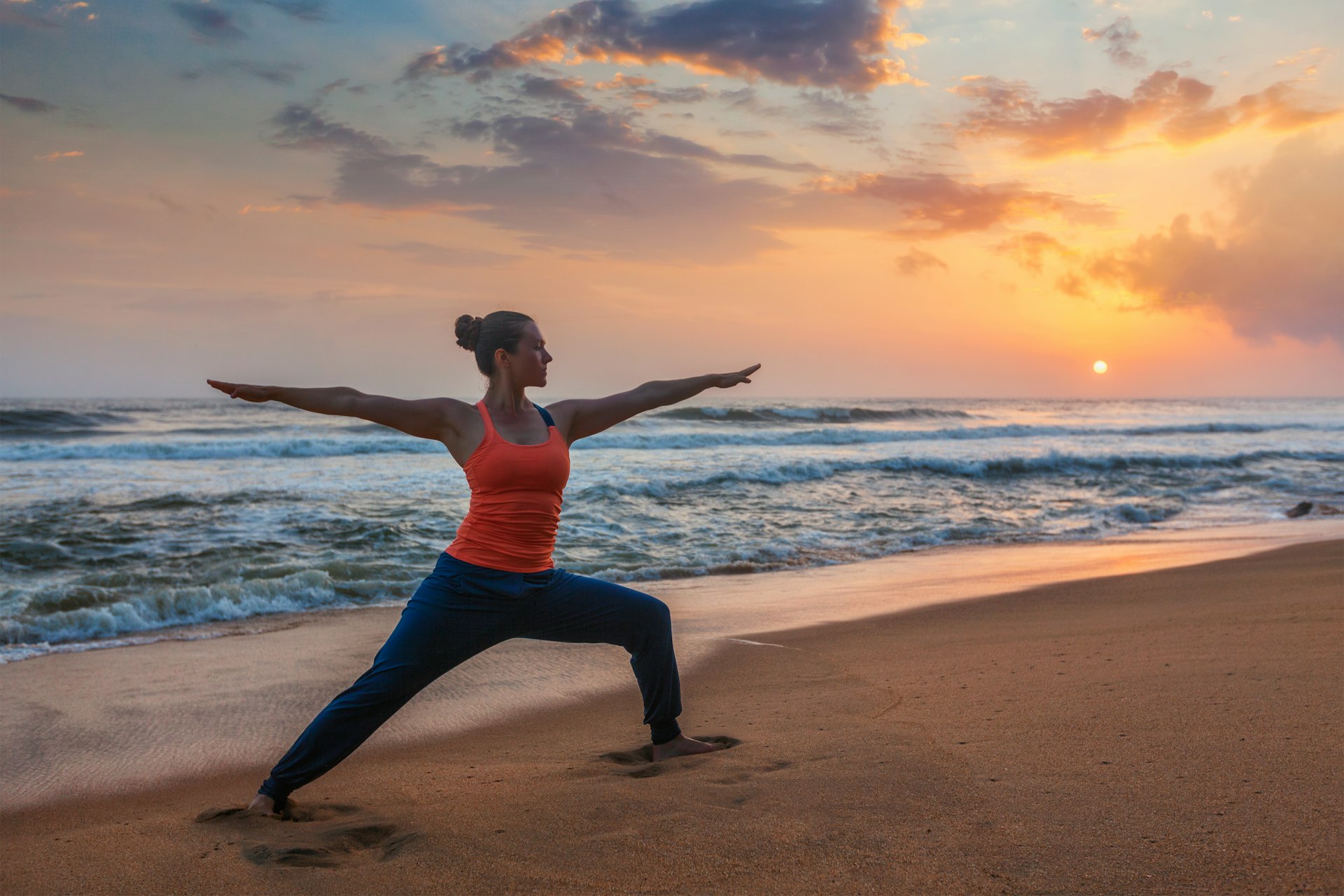 Woman doing a Virabhadrasana stance while practising Hatha yoga on a beach during sunset.