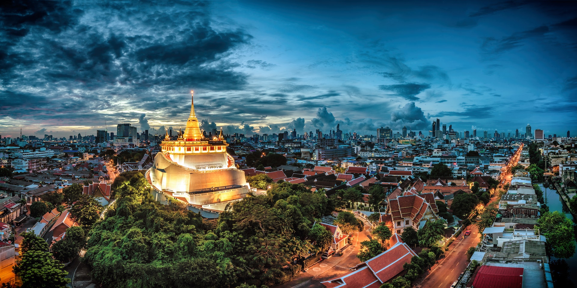 Wat Saket, The Golden Mount Temple in Bangkok, Thailand is lit up against a grey cloudy sky and the dark houses and skyscrapers of the city