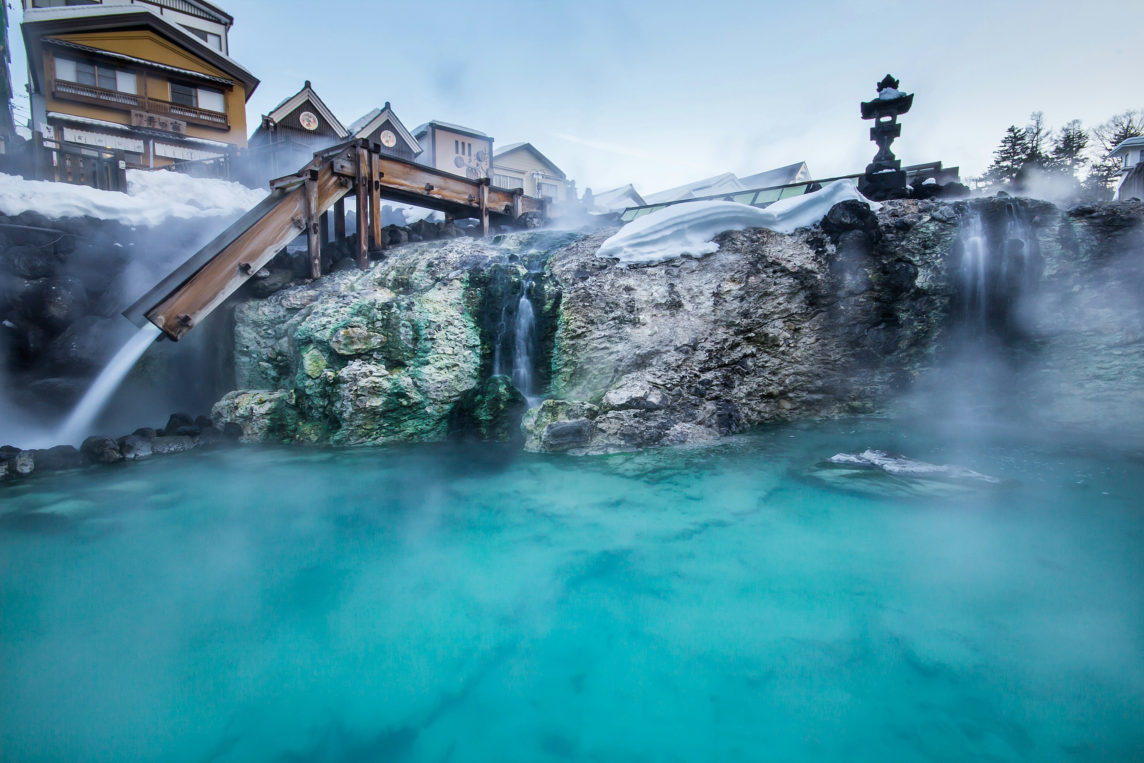 A pool of hot spring water is in the forground as steam rises from it and from the water flowing into it from a wooden trough.