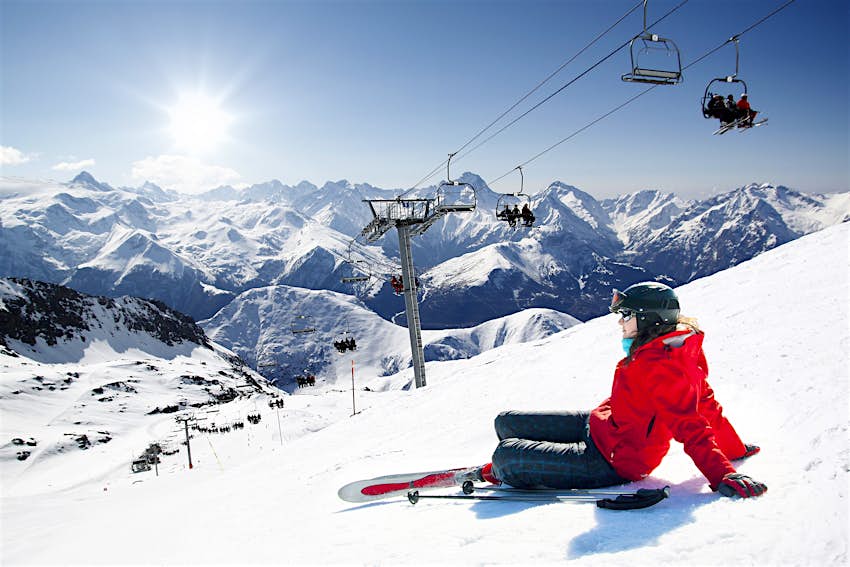 A skier wearing a red ski jacket and a ski helmet sits at the top of a slope near a cable car staring out at the snow-covered mountains