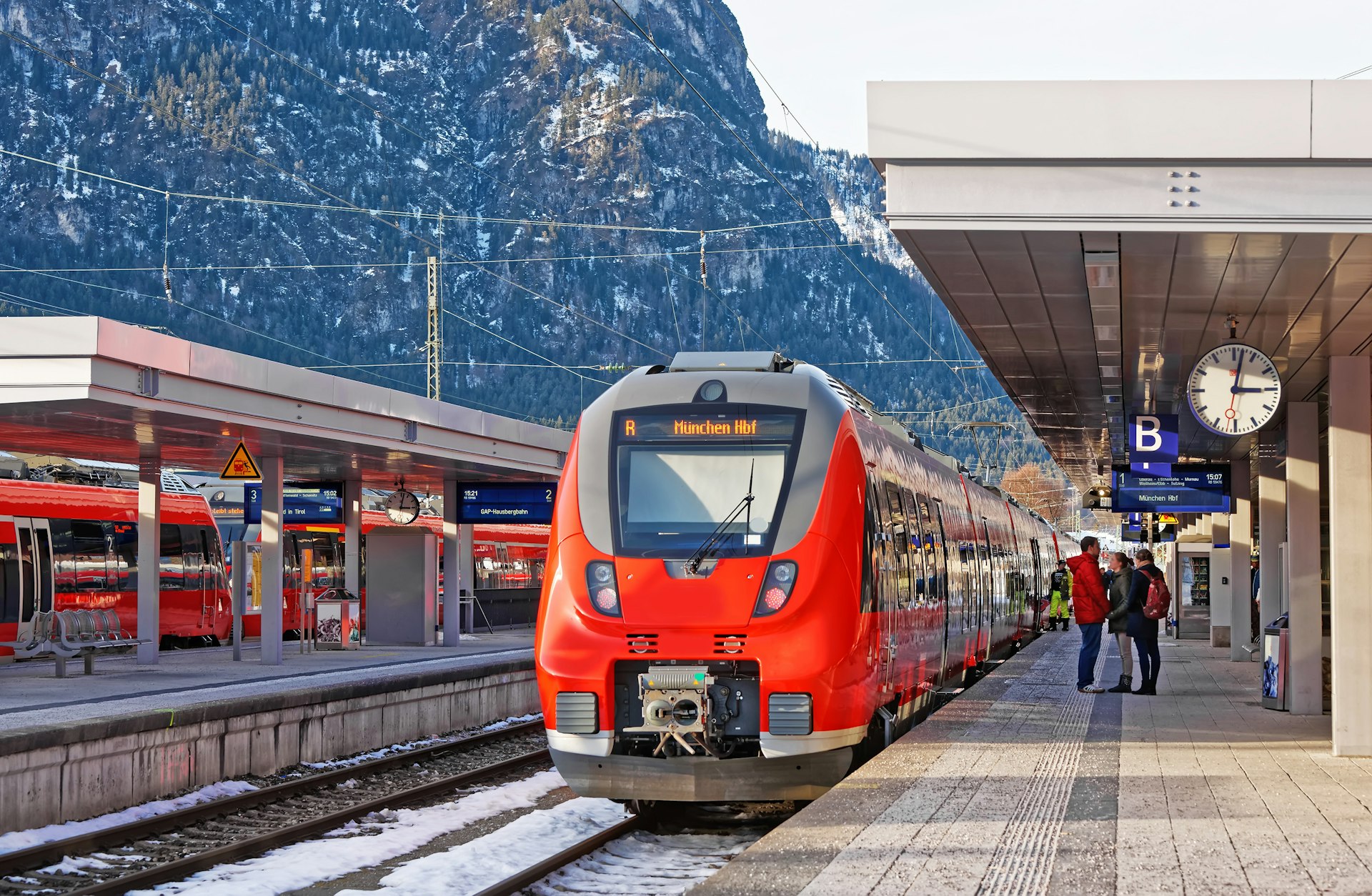 A red train standing at a platform at a station in Germany. A large black mountain rises in the background