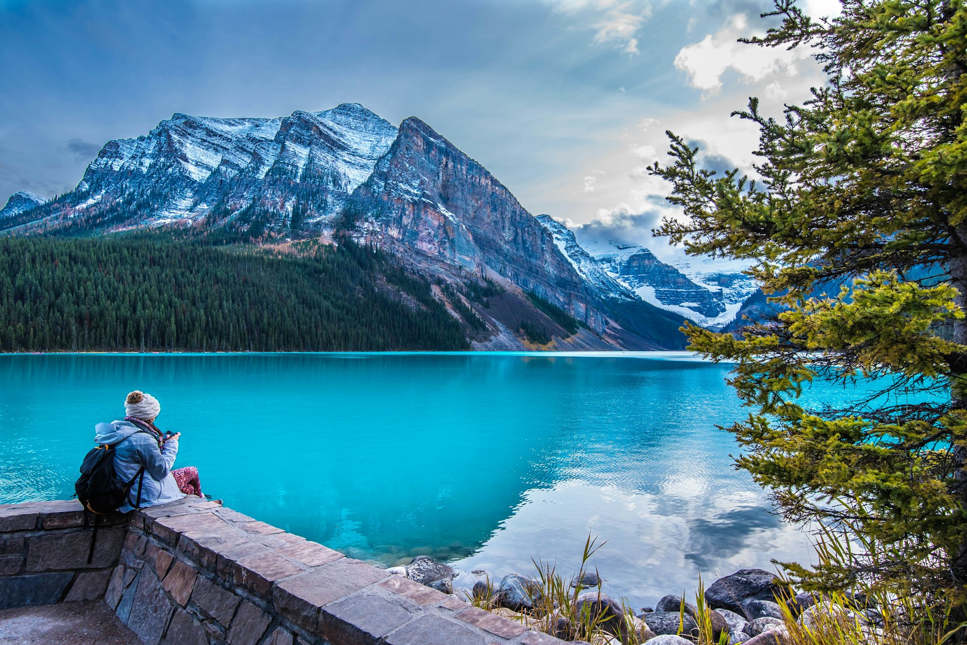 A woman sits on a wall looking out over a stunning turquoise lake surrounded by mountains