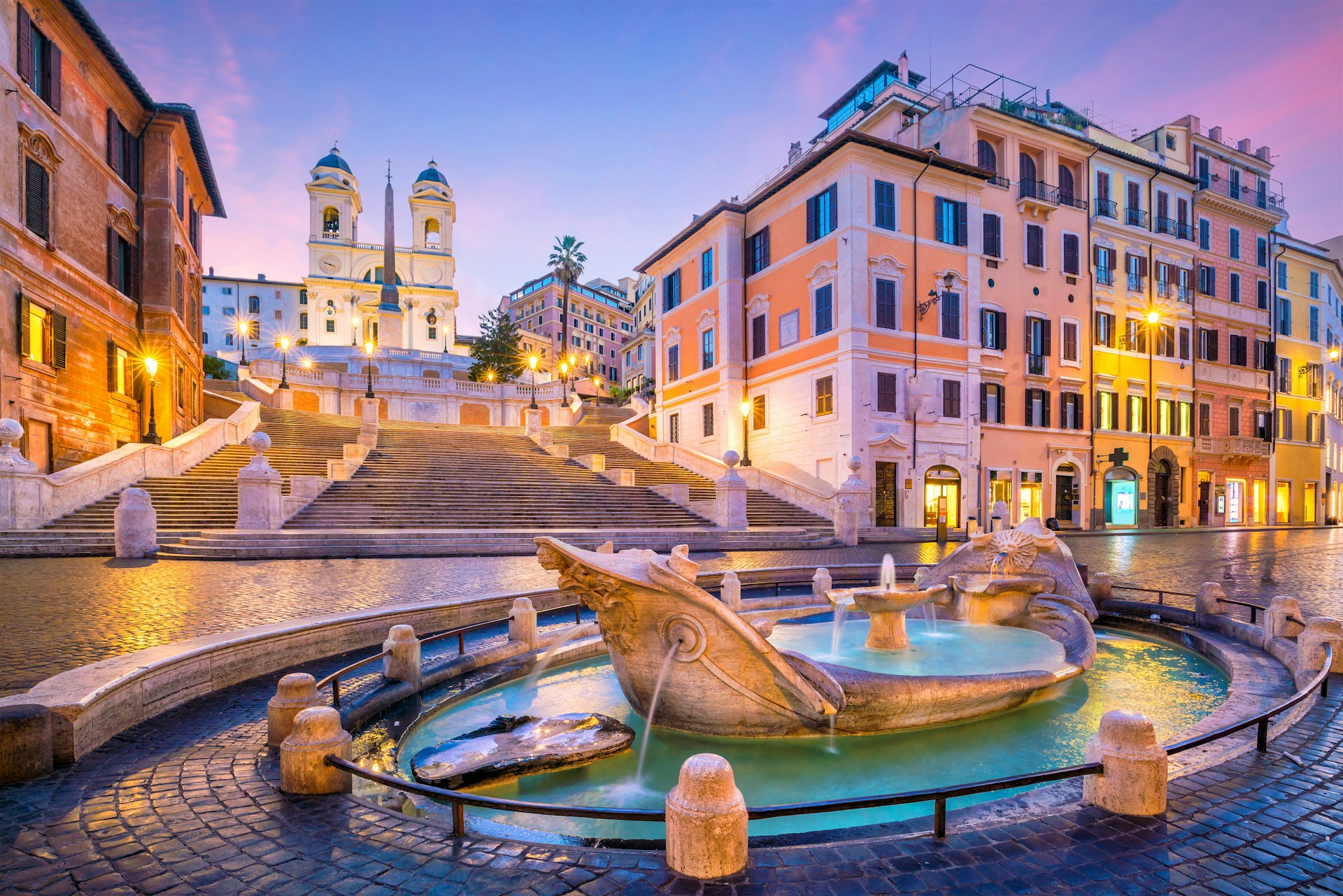 A series of steps lead down to a Roman plaza with cobbled stone streets and in the center of the photo is a marble foundation in the shape of a boat. 