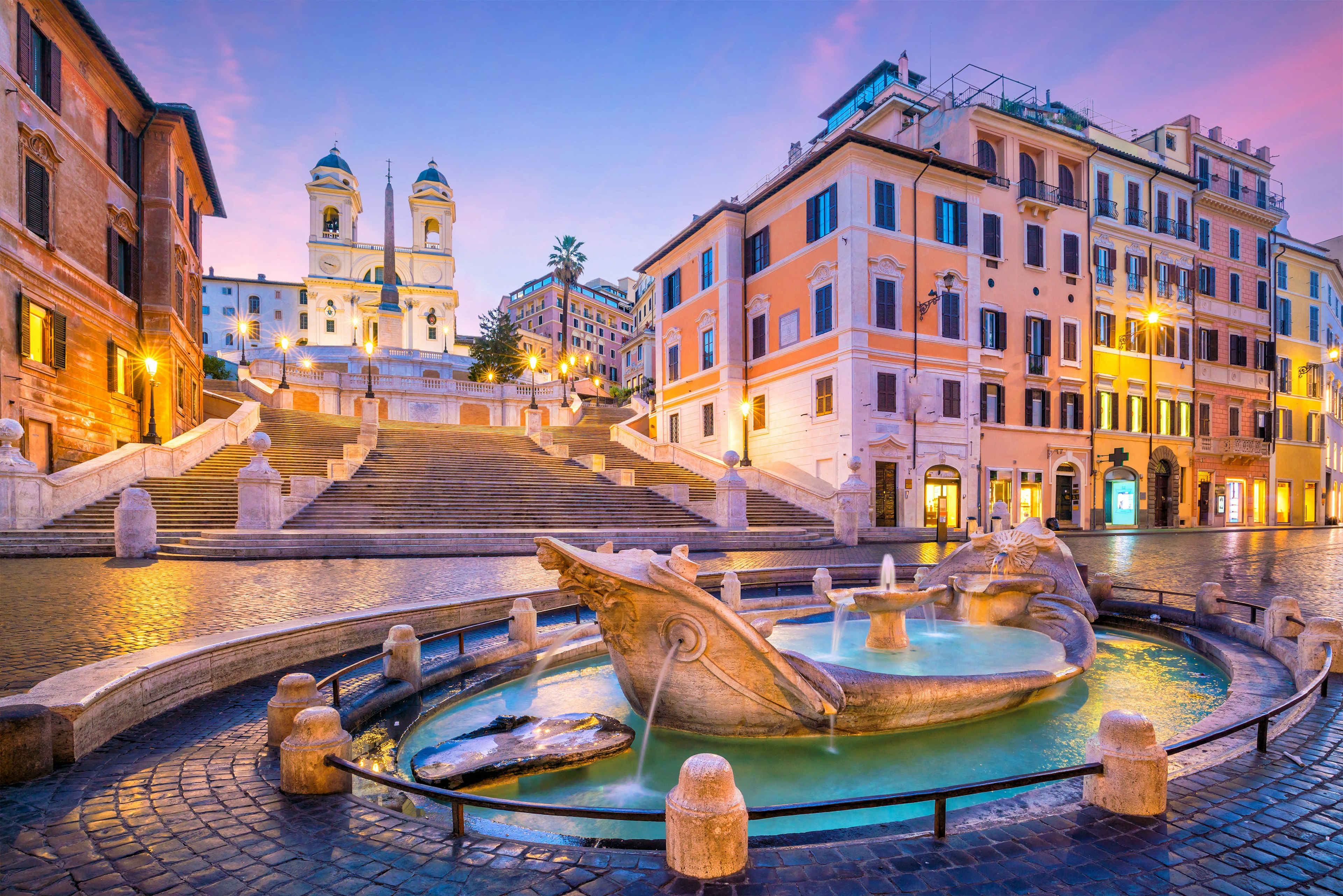 A series of steps lead down to a Roman plaza with cobbled stone streets and in the center of the photo is a marble foundation in the shape of a boat.