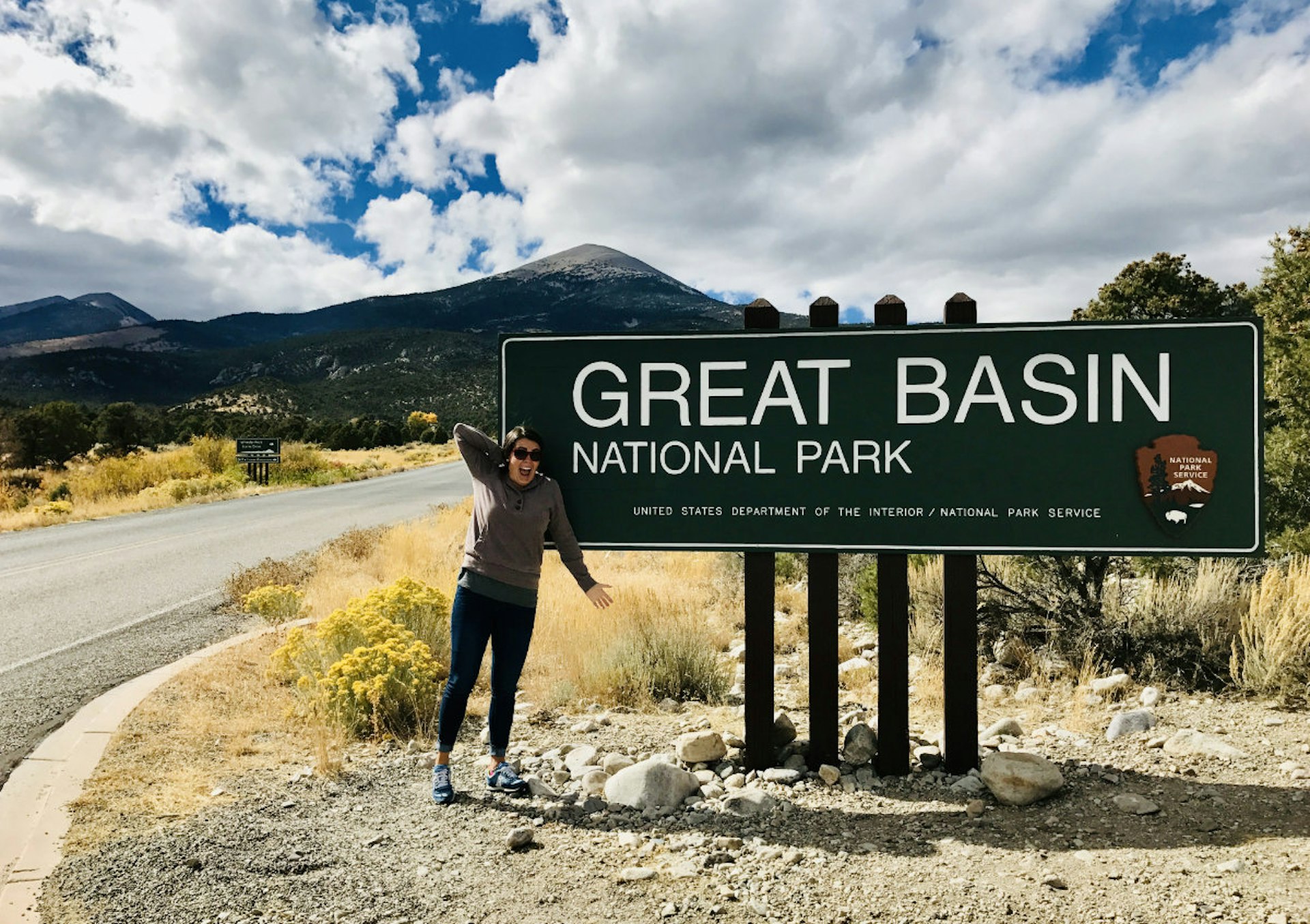 A large sign next to a deserted road stating "Great Basin National Park". A woman stands in front of the sign gesturing and smiling at the camera