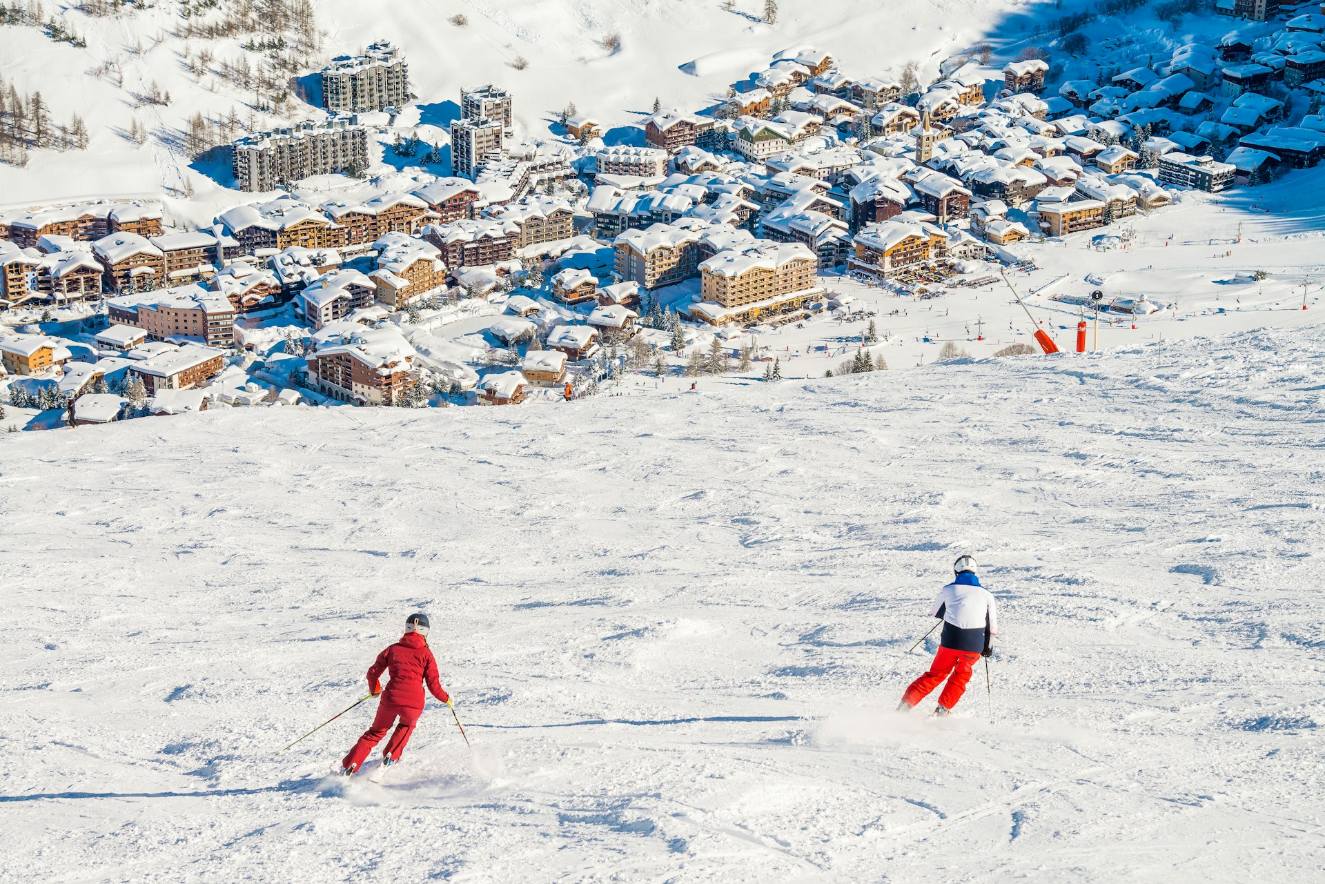 Two skiers viewed from behind as they descend a slope towards a town