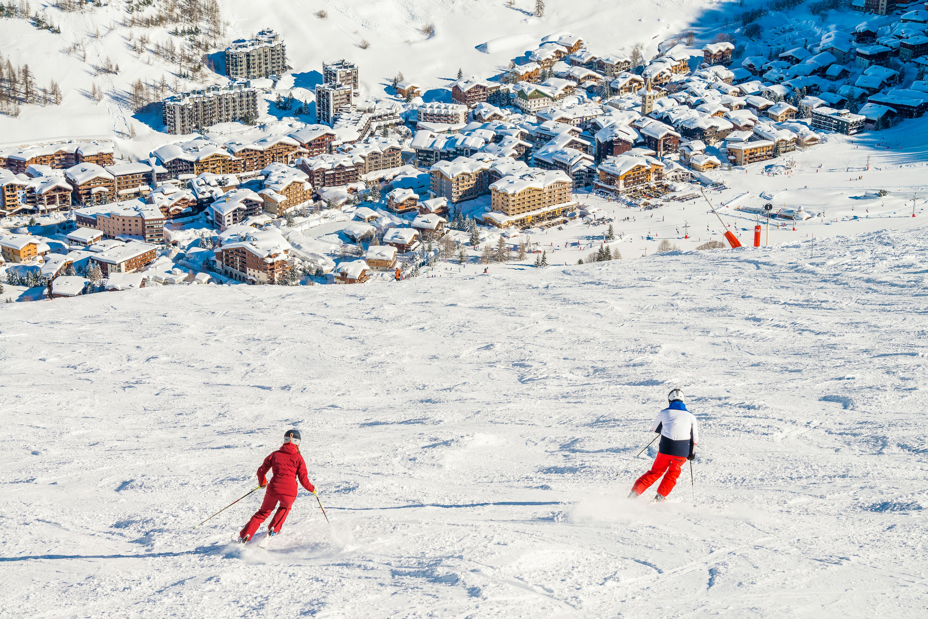 Two skiers viewed from behind as they descend a slope towards a town