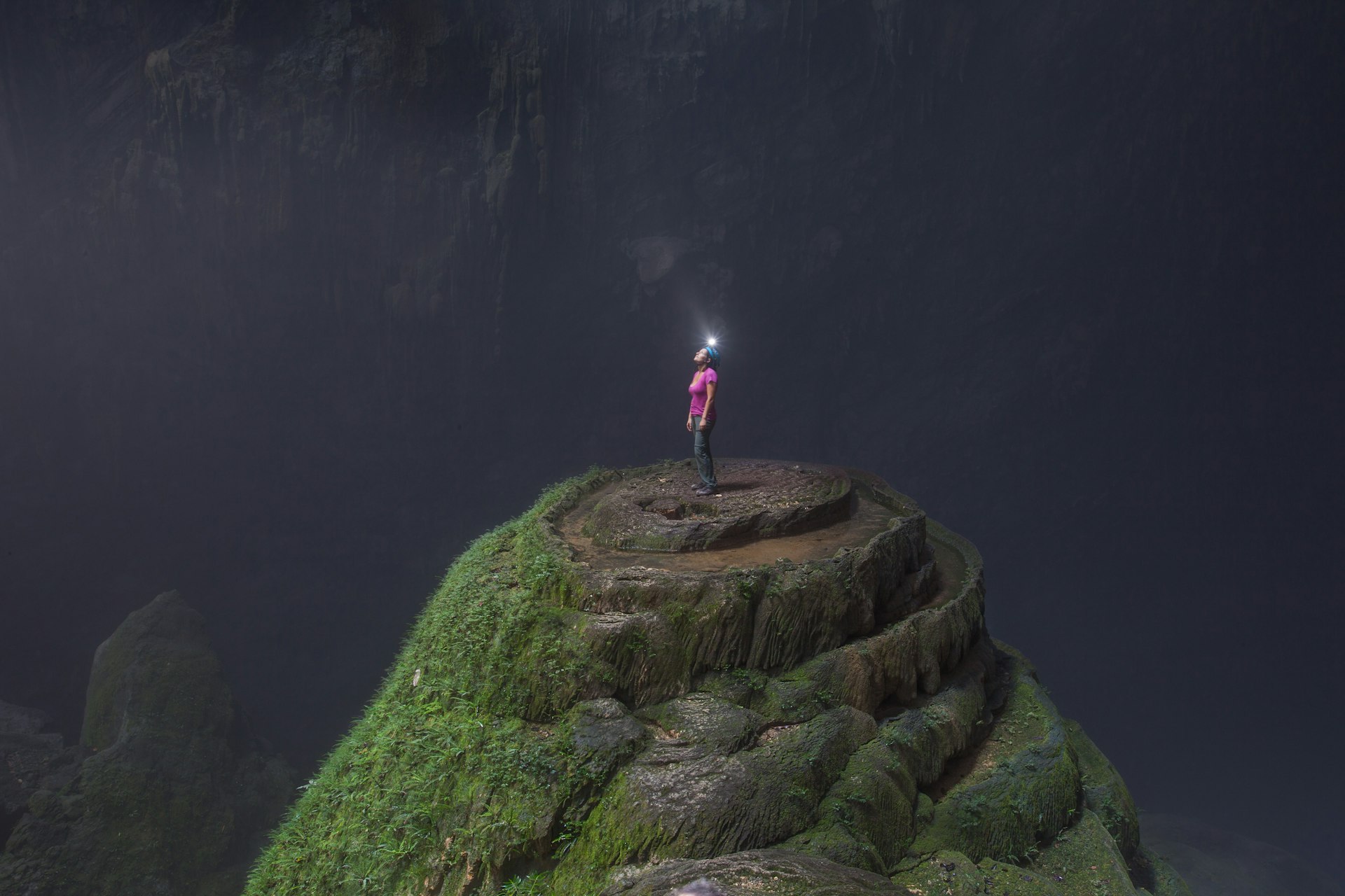 A woman wearing a helmet with a light stands at the top of a jagged rock gazes up to a hole in the ceiling in the expansive Hang Son Doong cave.