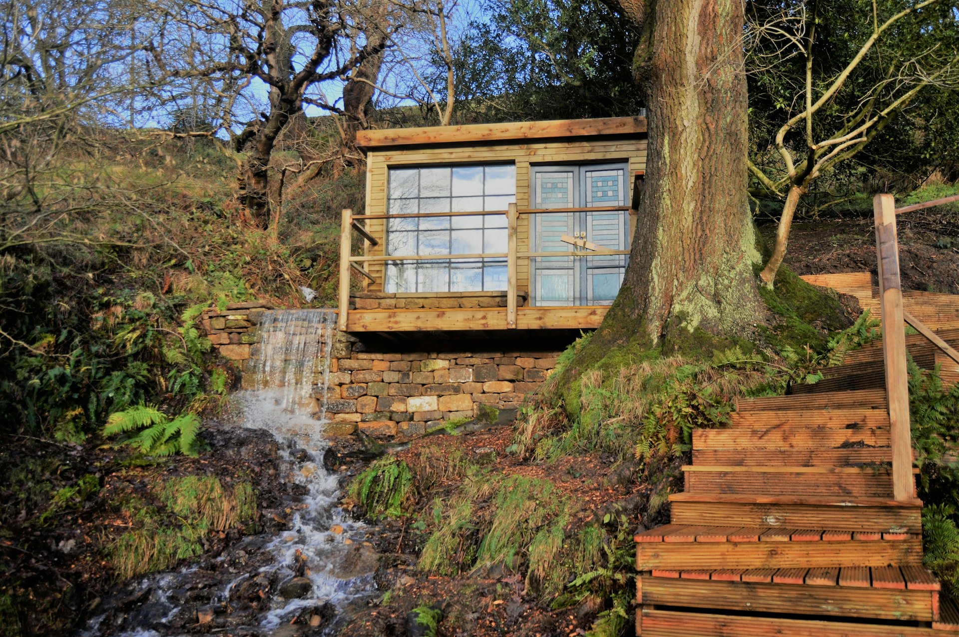 A cabin with large glass windows perches above a small waterfall