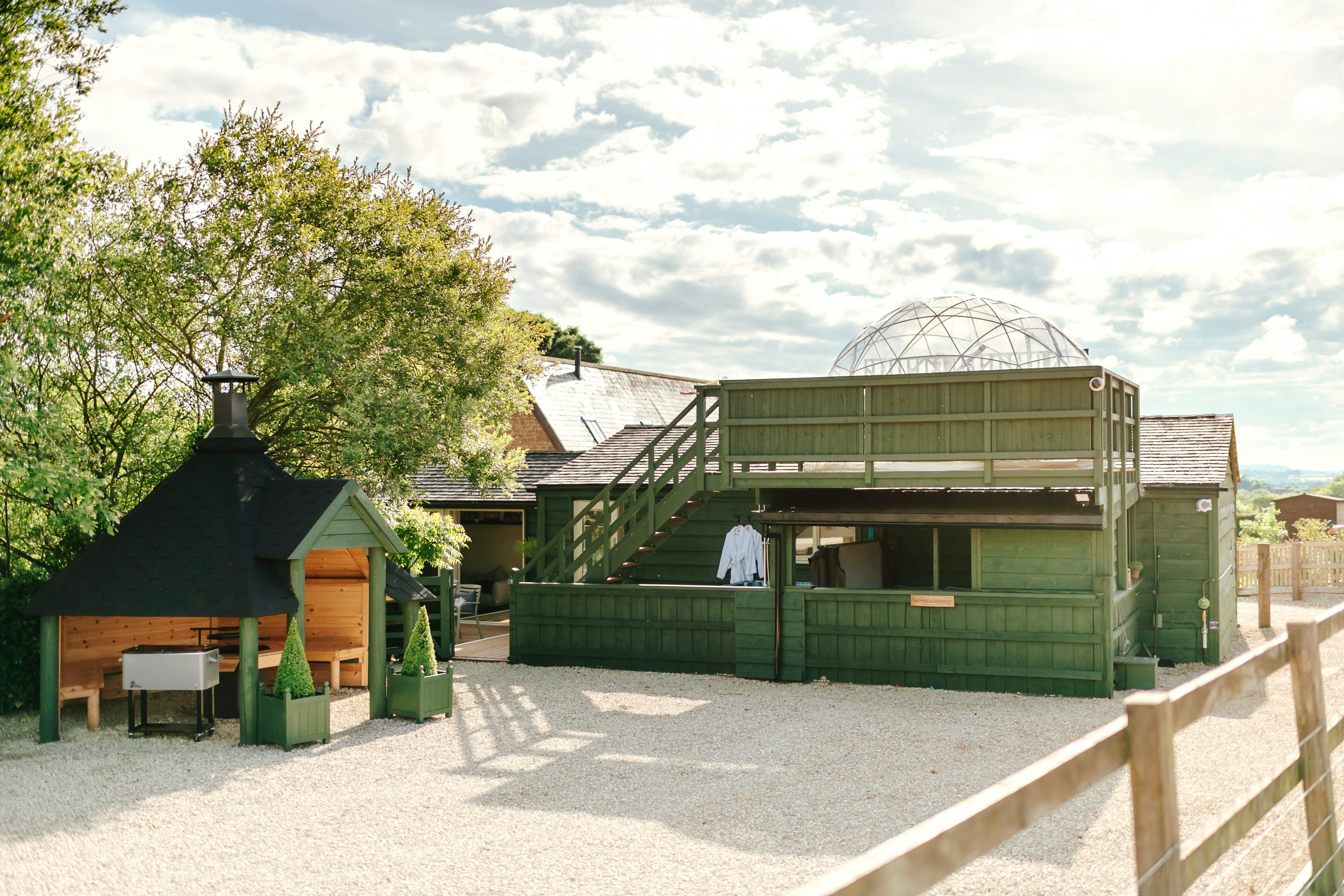 A wooden hut housing a pizza oven, and green wooden staircase leading to the roof of a cabin on a sunny day