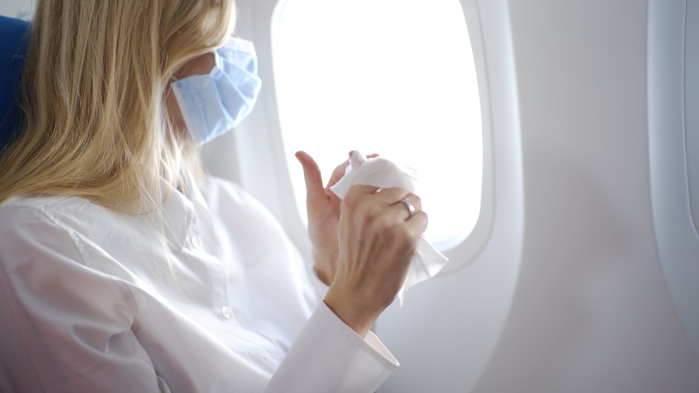 Portrait of young beautiful woman with long blond hair wearing mask and white shirt inside airplane and cleaning her hands using wet wipes.