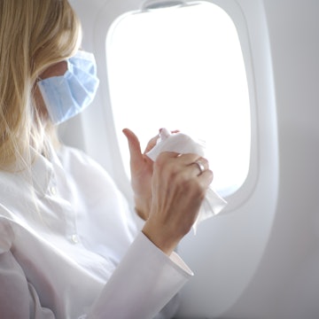 Portrait of young beautiful woman with long blond hair wearing mask and white shirt inside airplane and cleaning her hands using wet wipes.