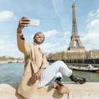 Woman taking a selfie while sitting on a bridge over the river Seine with the Eiffel tower in the background.
