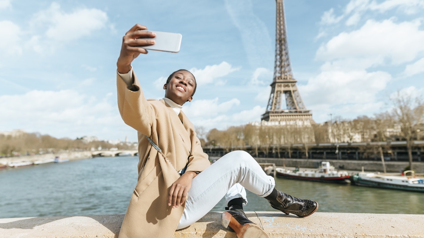 Woman taking a selfie while sitting on a bridge over the river Seine with the Eiffel tower in the background.