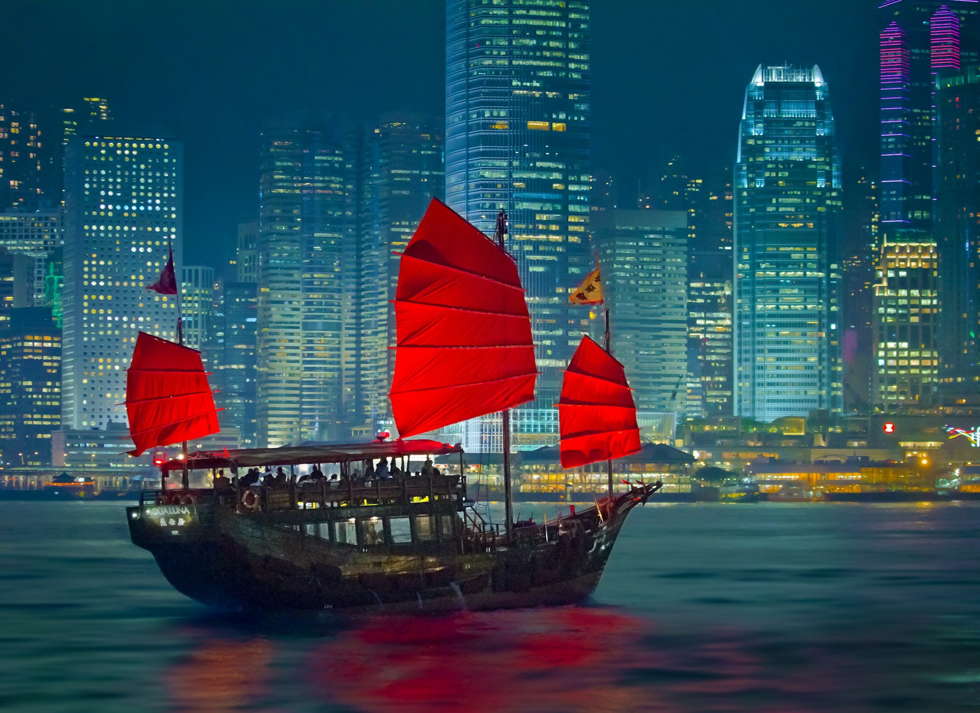 An old-fashioned junk boat with red sails in Hong Kong harbor, contrasts with the glass-and-steel skyscrapers in the background