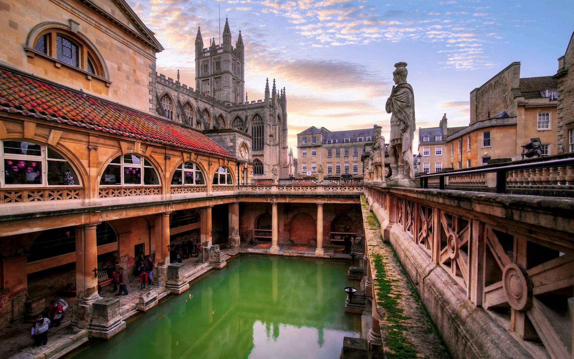 An open-air rectangular Roman bath filled with green water surrounded by a colonnaded walkway lined with statues 