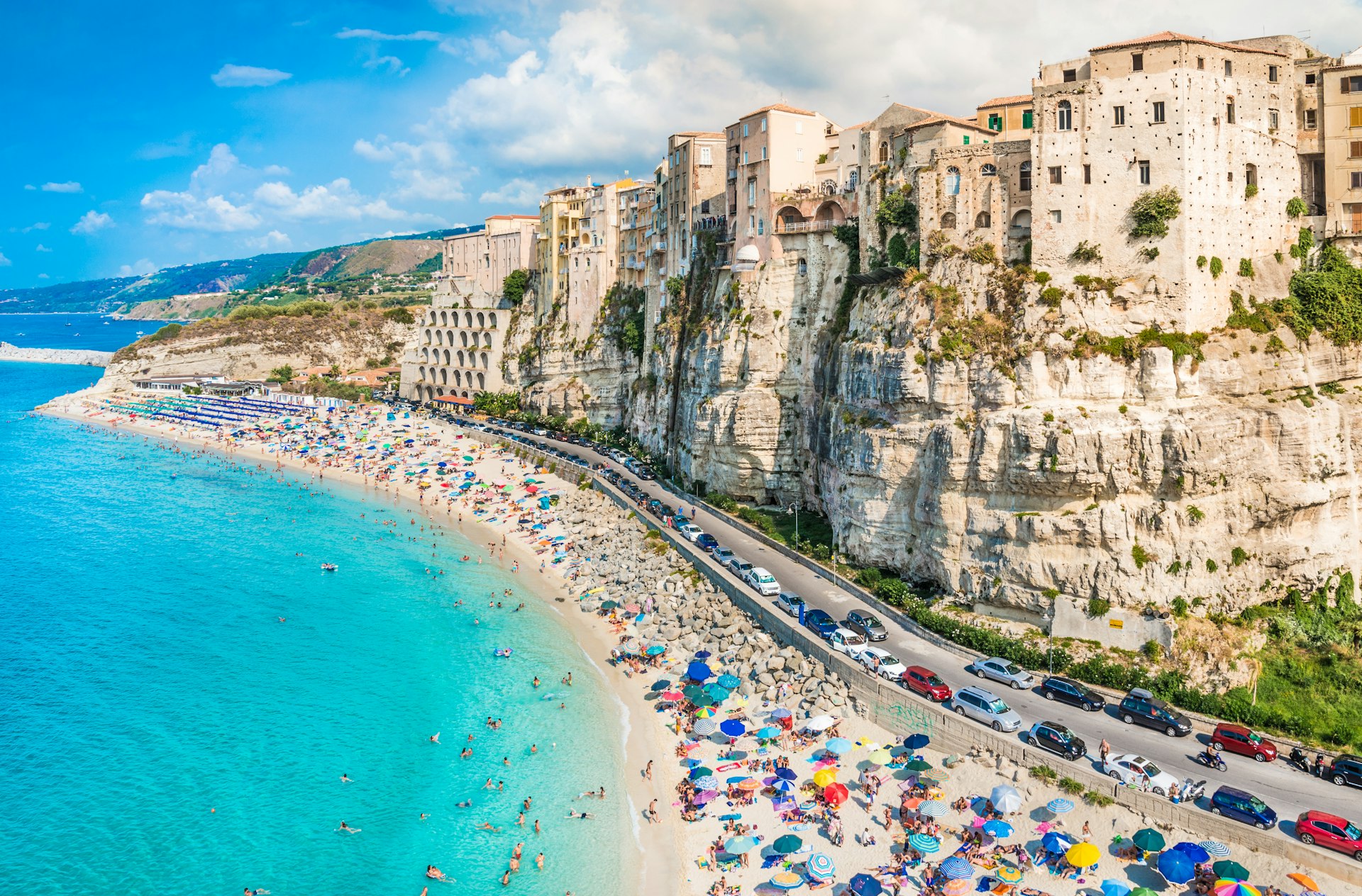 A high-angled view of Tropea Beach in Calabria, Italy. A sweep of golden white sand, which is full of sun bathers, is lapped by light clue waters. Behind it are a series of white cliffs.