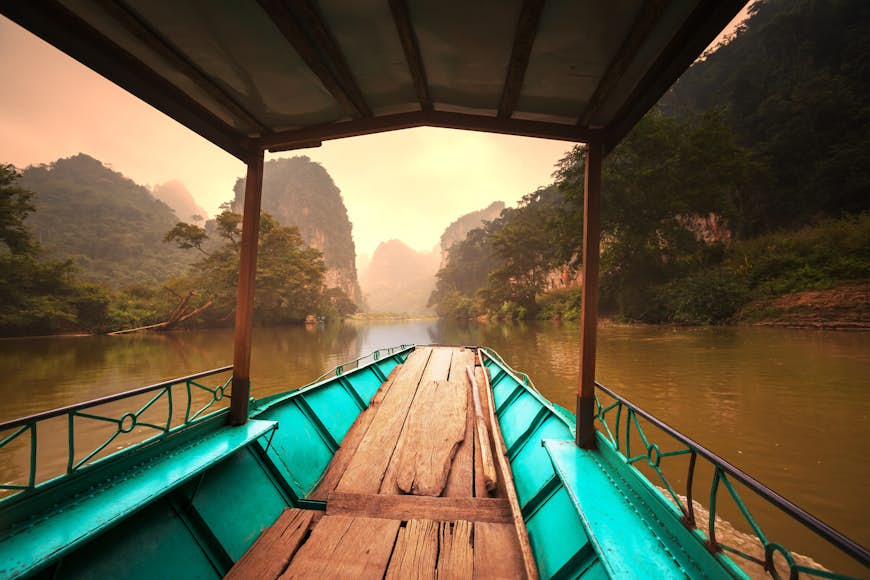 An empty boat is tied up on the banks of a lake in Ba Be National Park. Beyond the boat the shimmering still waters of the lake is visible, which is backed by numerous mountains, most of which are covered in forest.