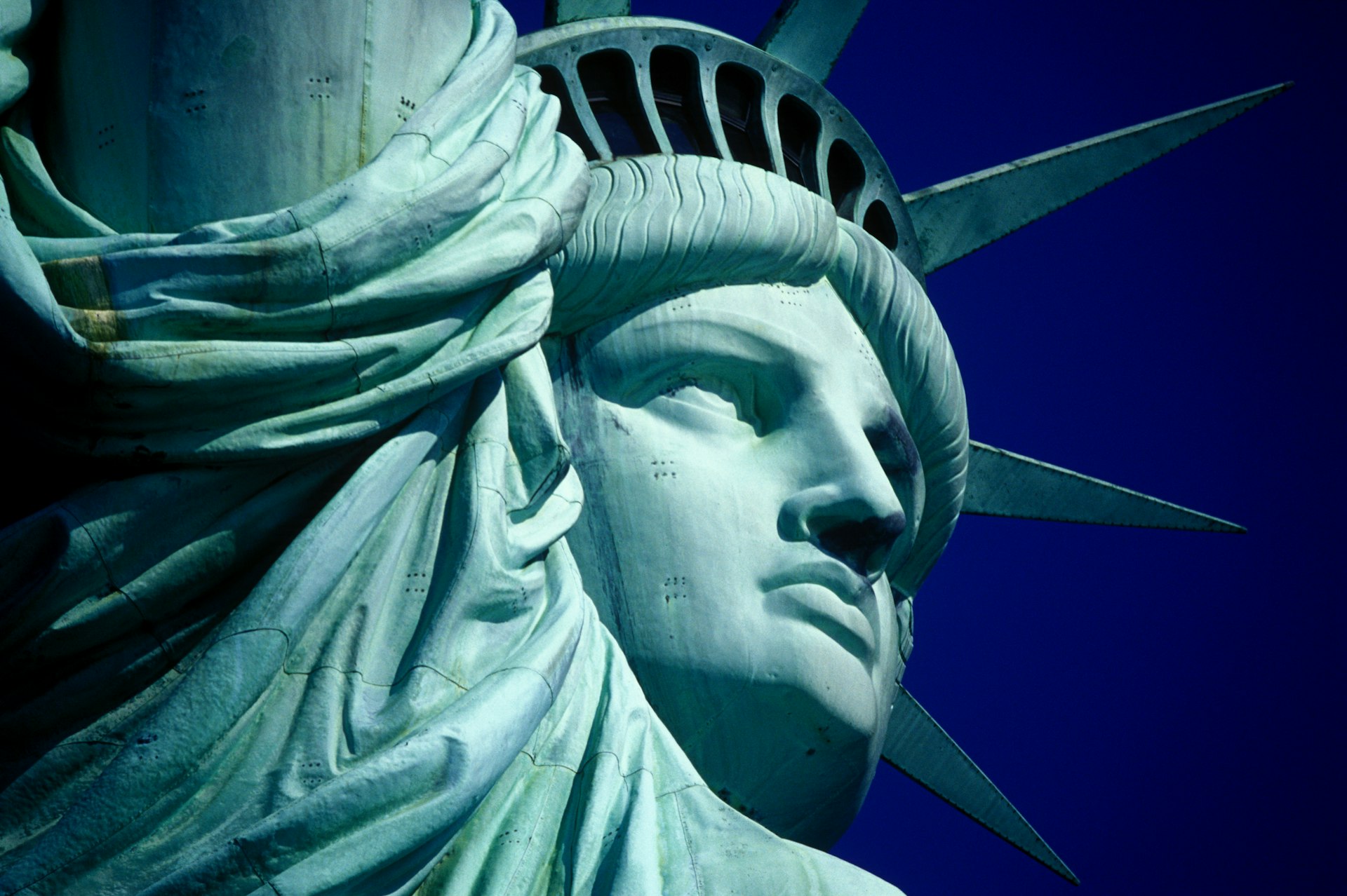 Cropped, up close photo of the Statue of Liberty's face and crown cut against a clear blue sky