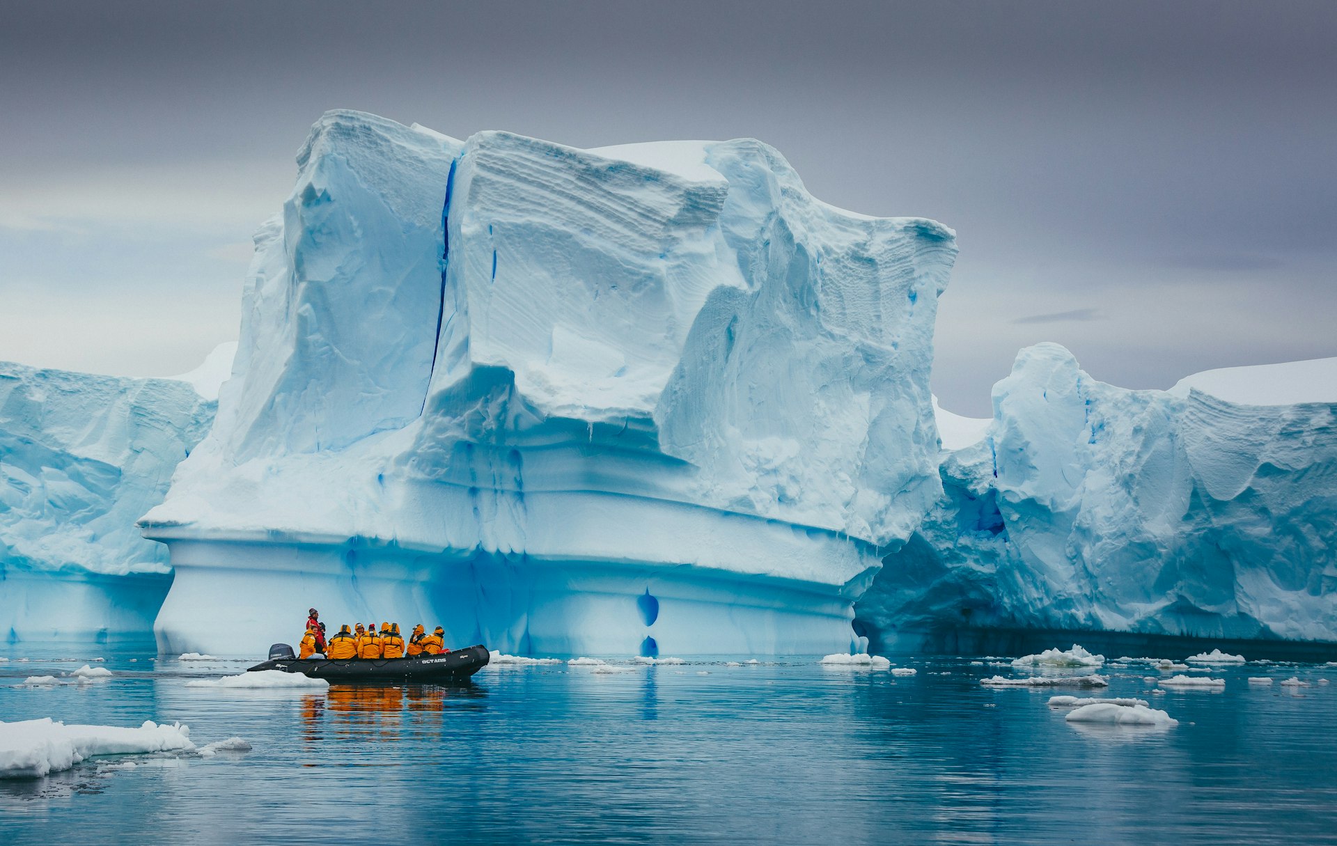 A small boat loaded with people in yellow coats pulls up alongside a large blue-white iceberg