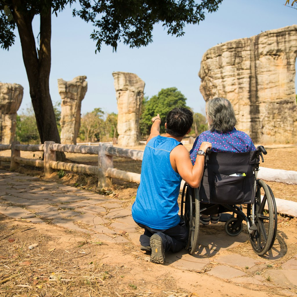 A man wearing blue singlet and black sport pants takes his old mother on wheelchair to see the strange rock formation of Mo Hin Khao (Thailand's Stonehenge) in Chaiyaphum Province, Thailand.