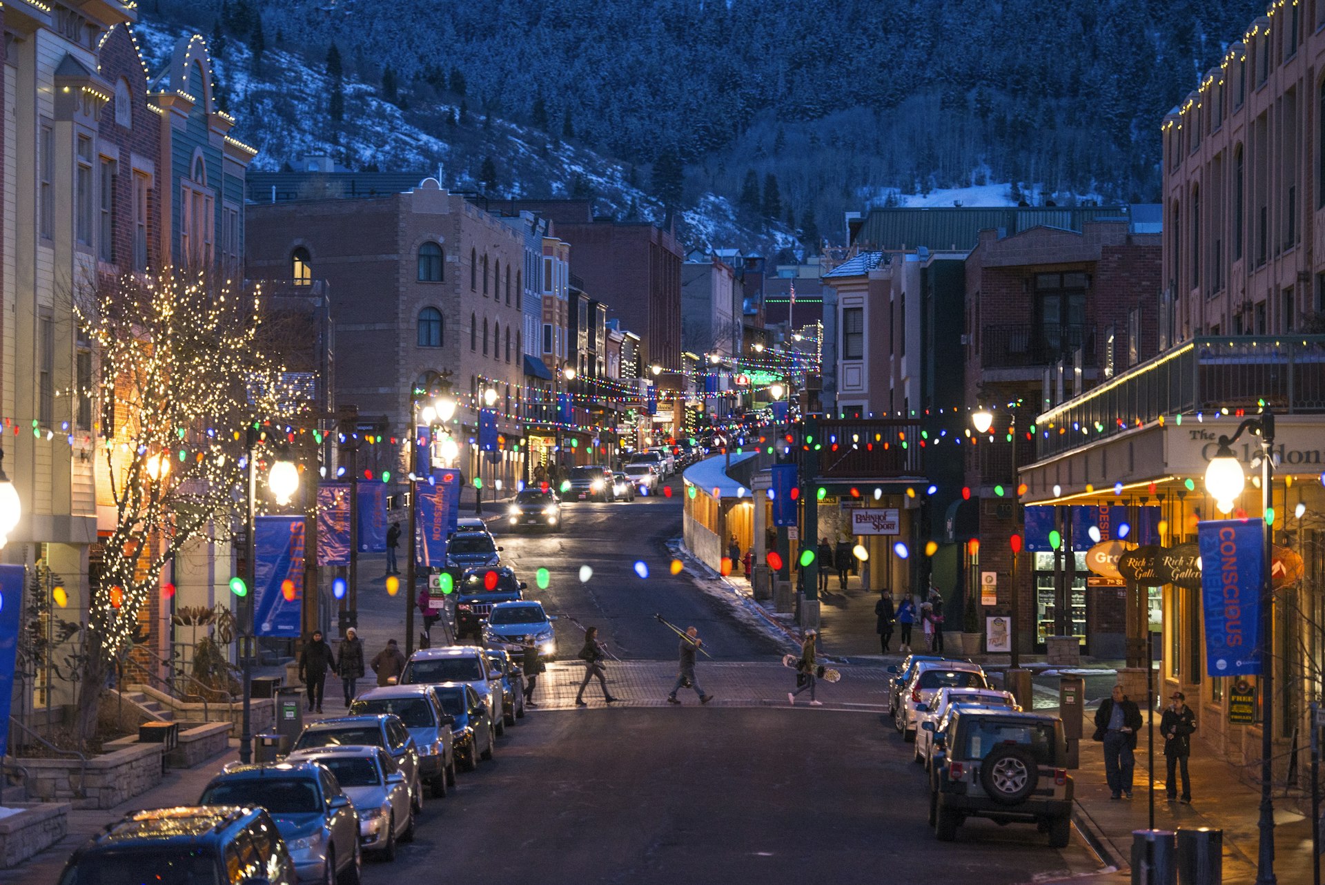 Skiers and a snowboarder cross a festively lit and decorated Main Street in Park City at night.
