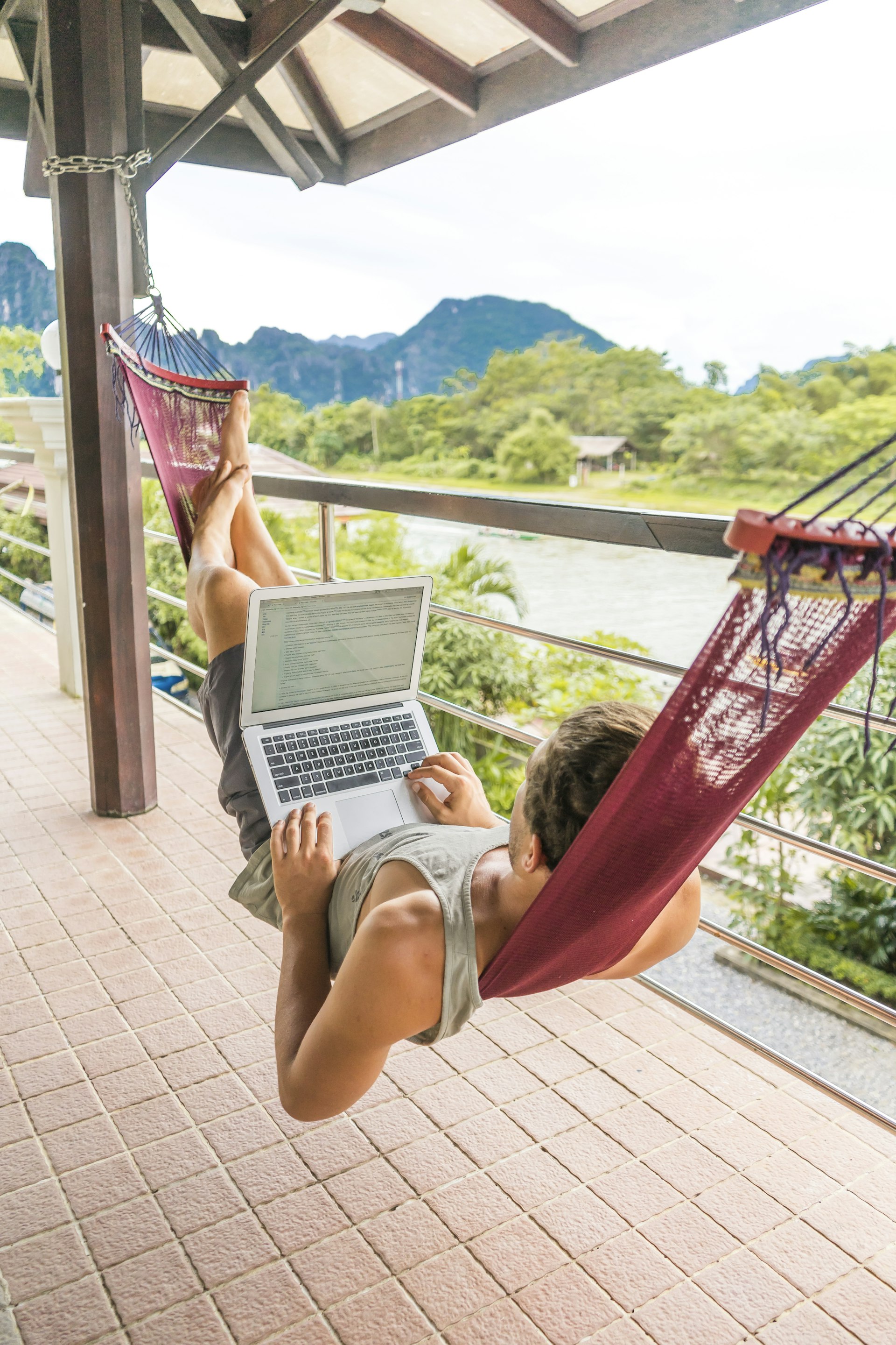 A young man lying in hammock that hangs on a balcony near a river. A laptop is balanced on his legs