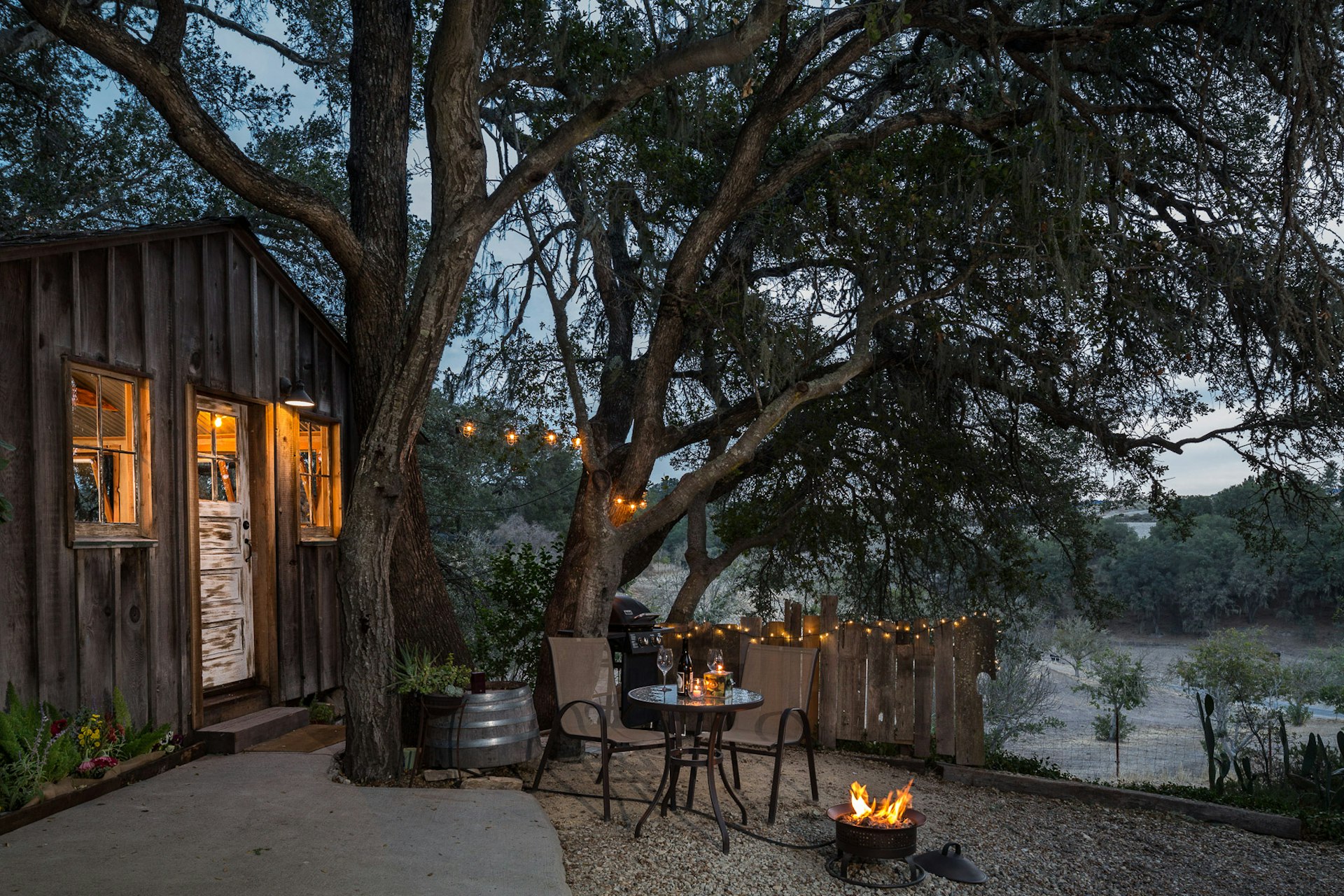 A cabin at dusk, lit from within, with a small bistro table, a firepit, and a fence with twinkle lights
