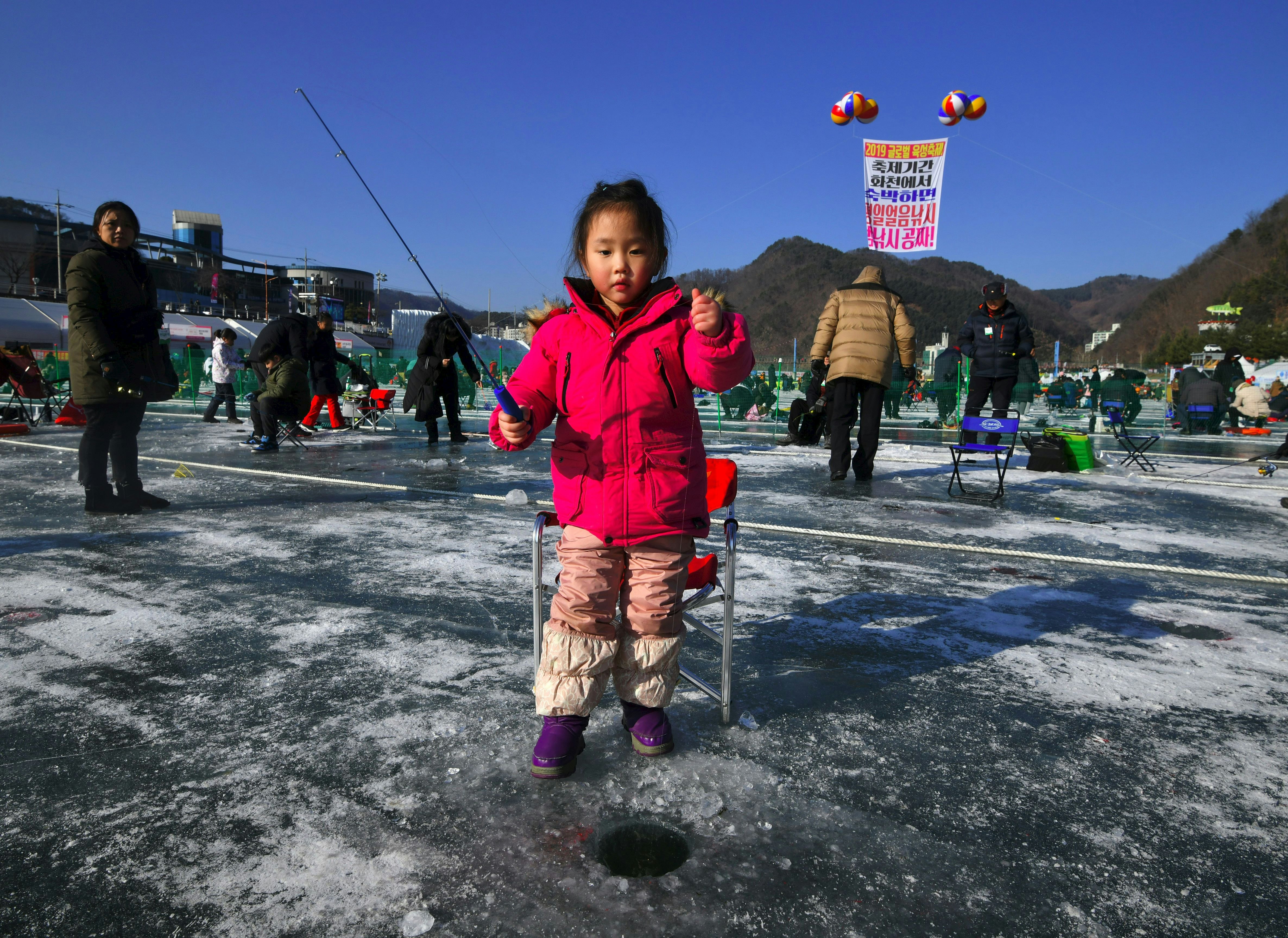 A little girl wearing a pink coat, pink winter pants and purple boots holds a fishing rod as she ice fishes during the Hwacheon Sanchenoeo Festival in South Korea. In the background a number of other people are also ice fishing. 