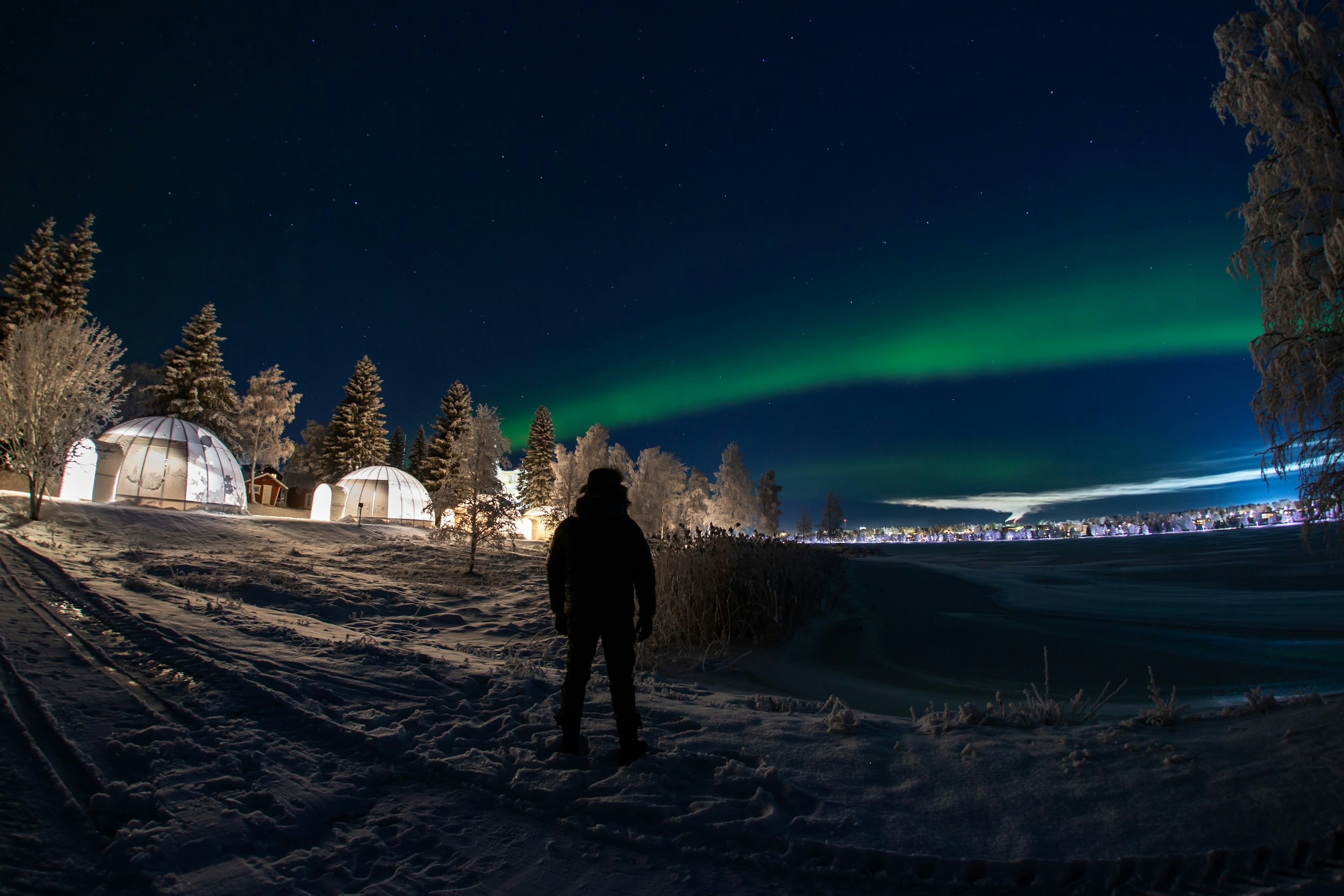 A man walking in front of igloo bubble cabins with the Northern Lights behind him