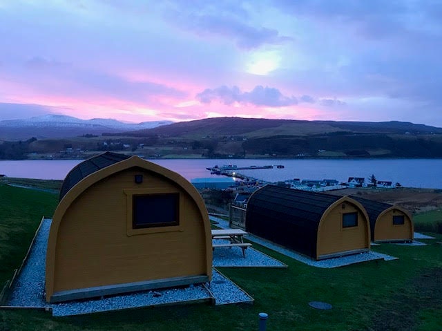 The back of three tiny wooden cabins facing a body of water with the sun rising beyond the hills