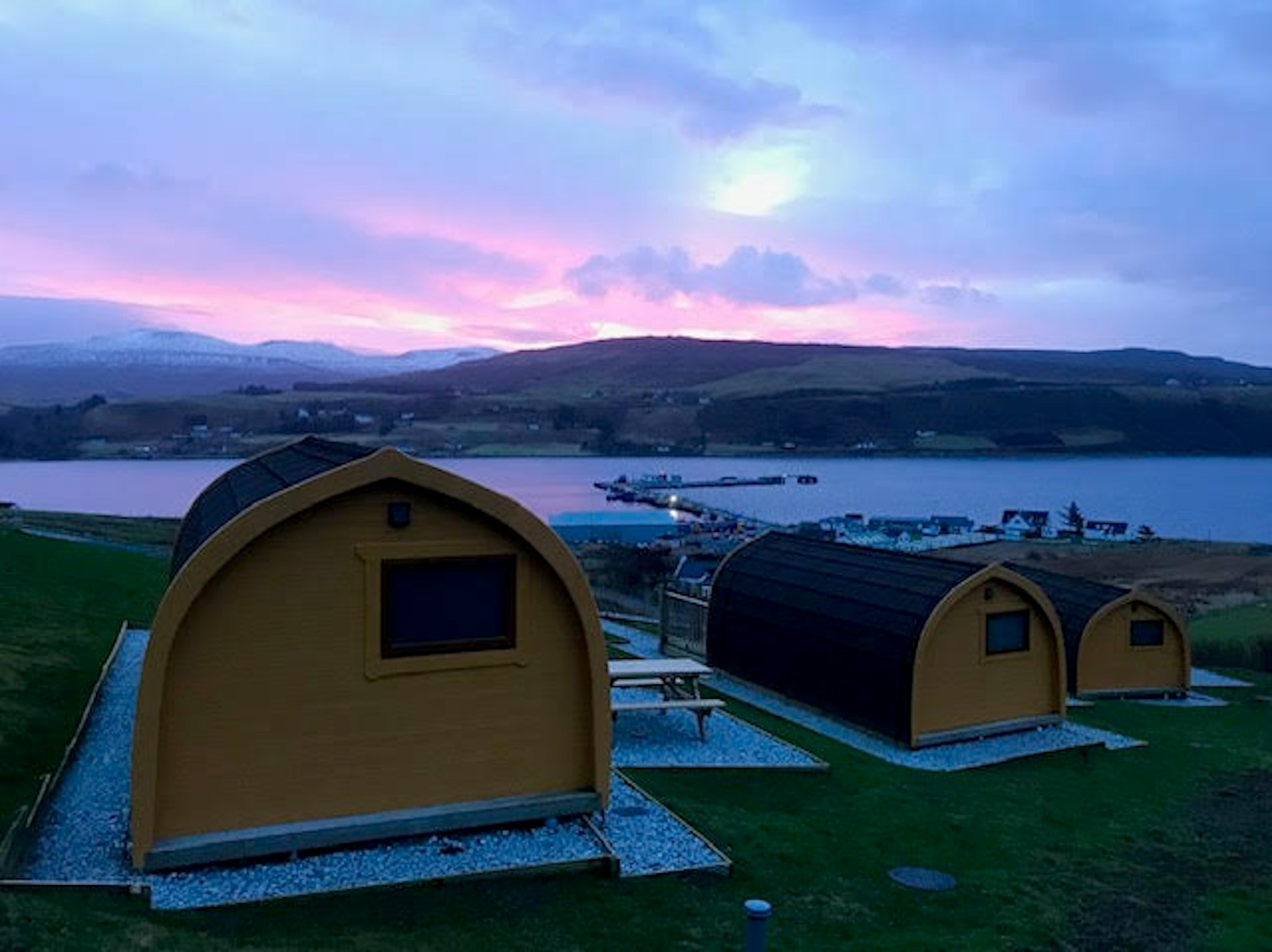 The back of three tiny wooden cabins facing a body of water with the sun rising beyond the hills